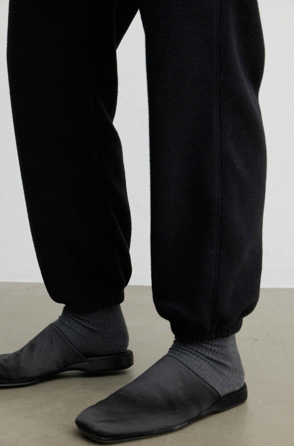 Close-up of a person's lower legs wearing black jogger sweatpants, dark gray ribbed socks, and black slip-on shoes on a light gray floor against a white wall.