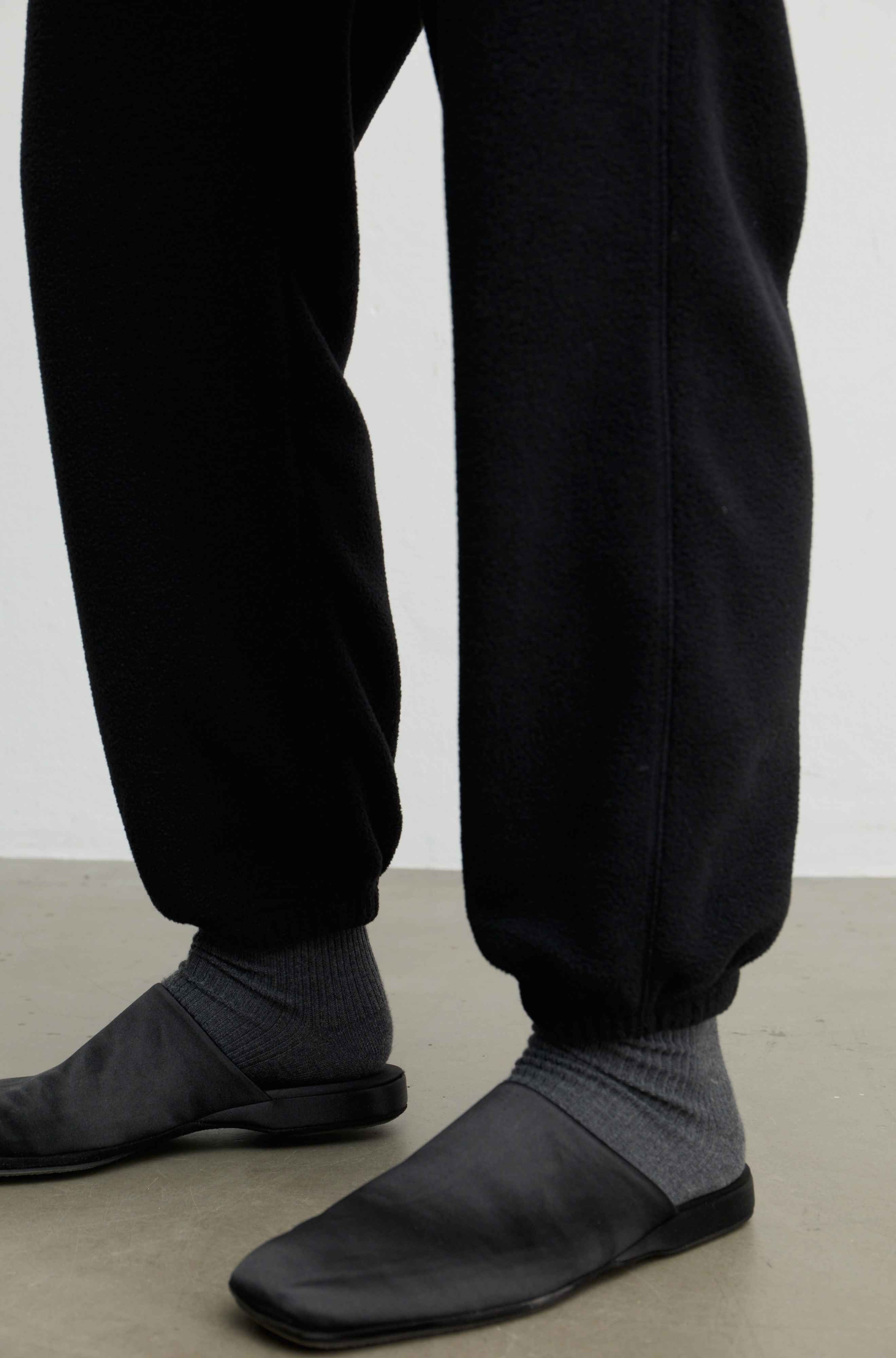 Close-up of a person's lower legs wearing black jogger sweatpants, dark gray ribbed socks, and black slip-on shoes on a light gray floor against a white wall.