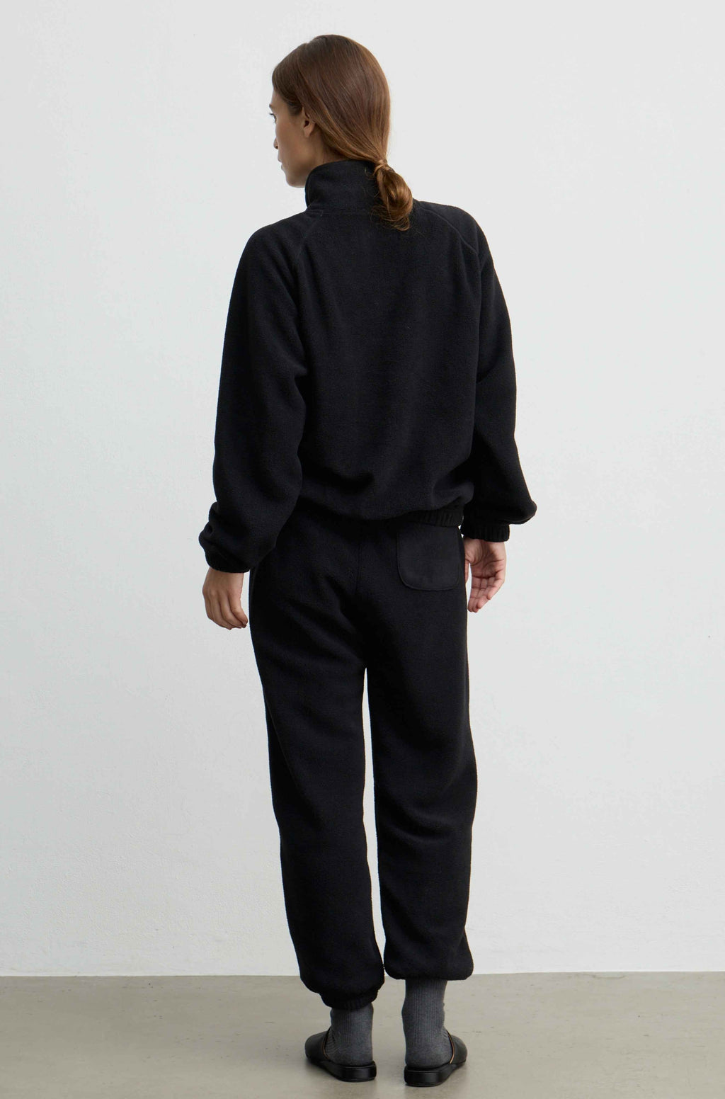 A model viewed from behind wearing a black fleece zip-up jacket and matching black fleece pants, standing against a plain white studio wall.