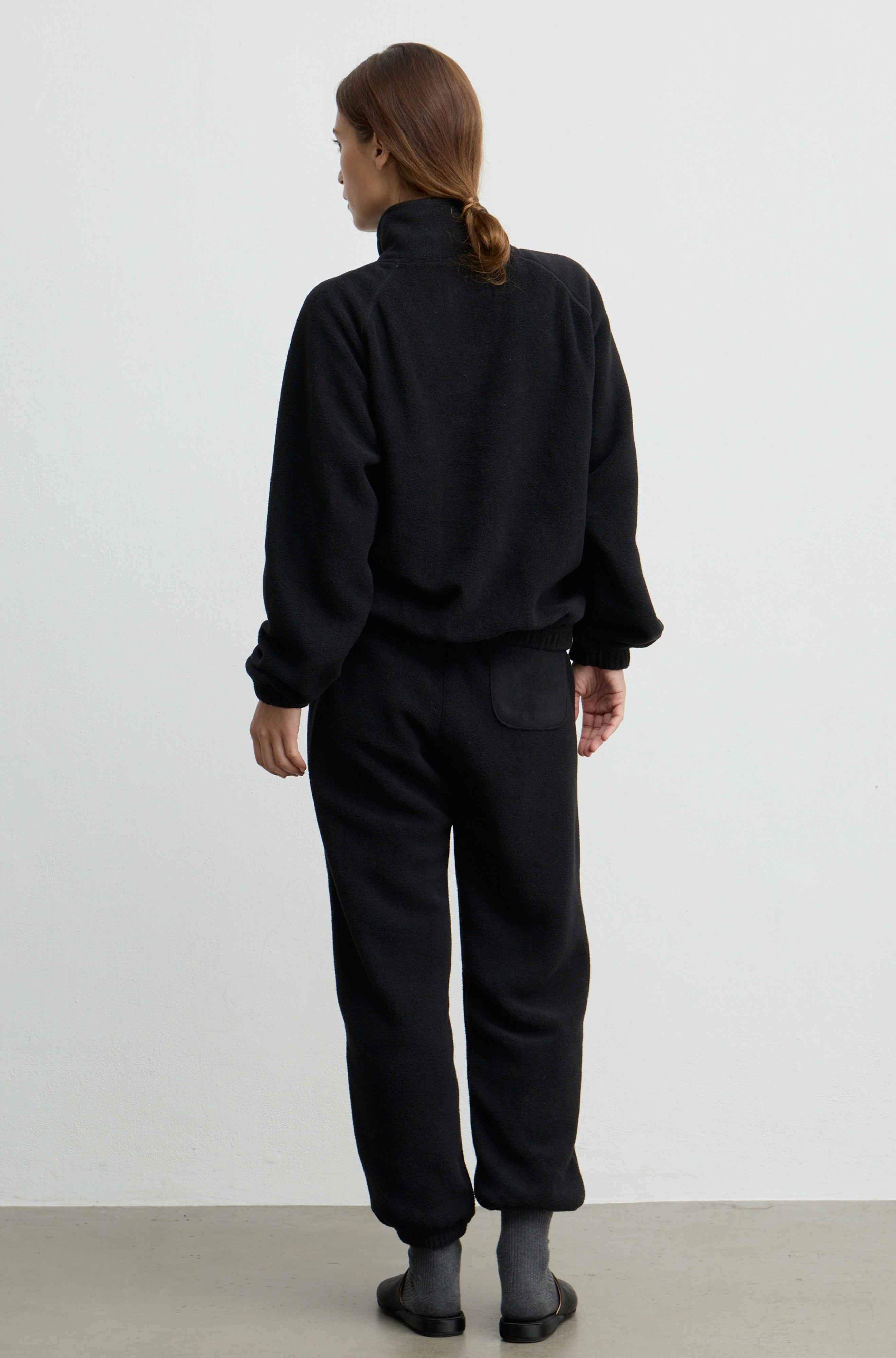 A model viewed from behind wearing a black fleece zip-up jacket and matching black fleece pants, standing against a plain white studio wall.