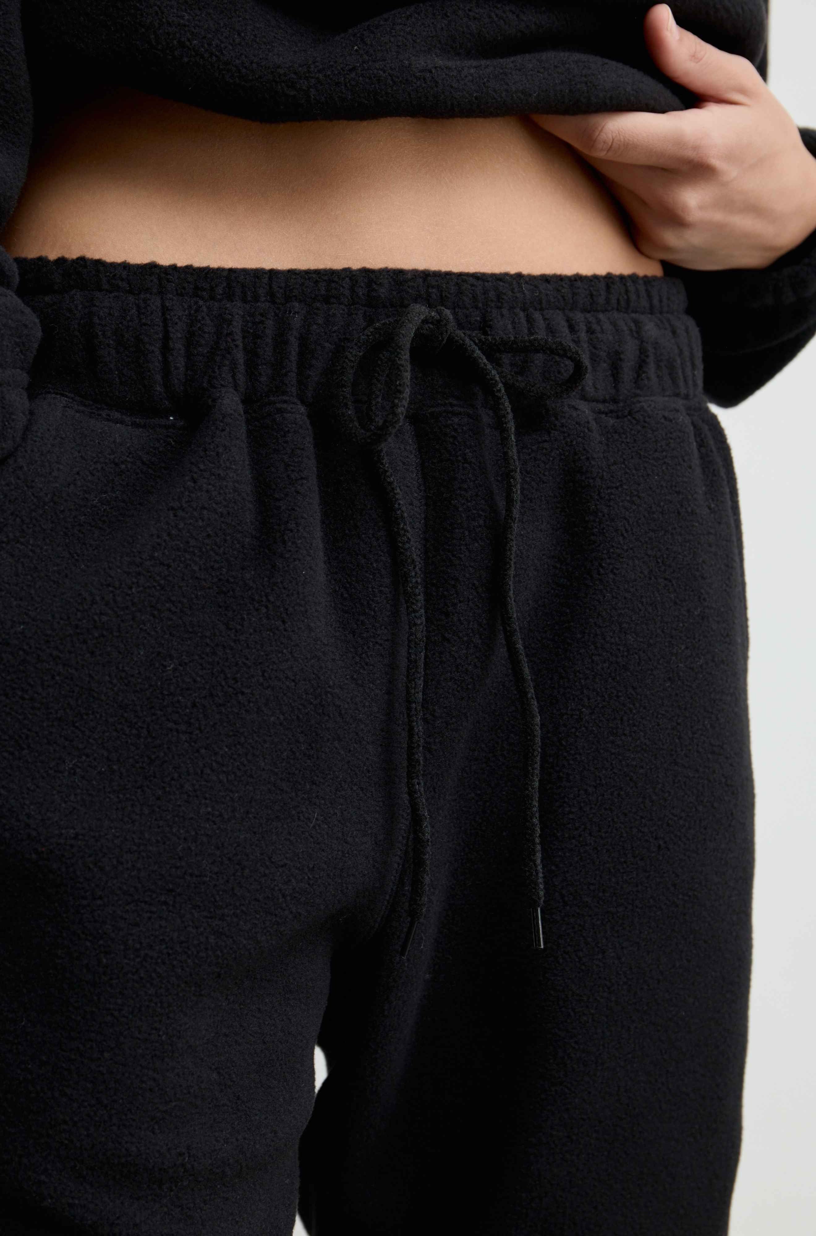 Close-up of a person wearing black fleece drawstring sweatpants, with the hand lifting a black top to show the waist area.
