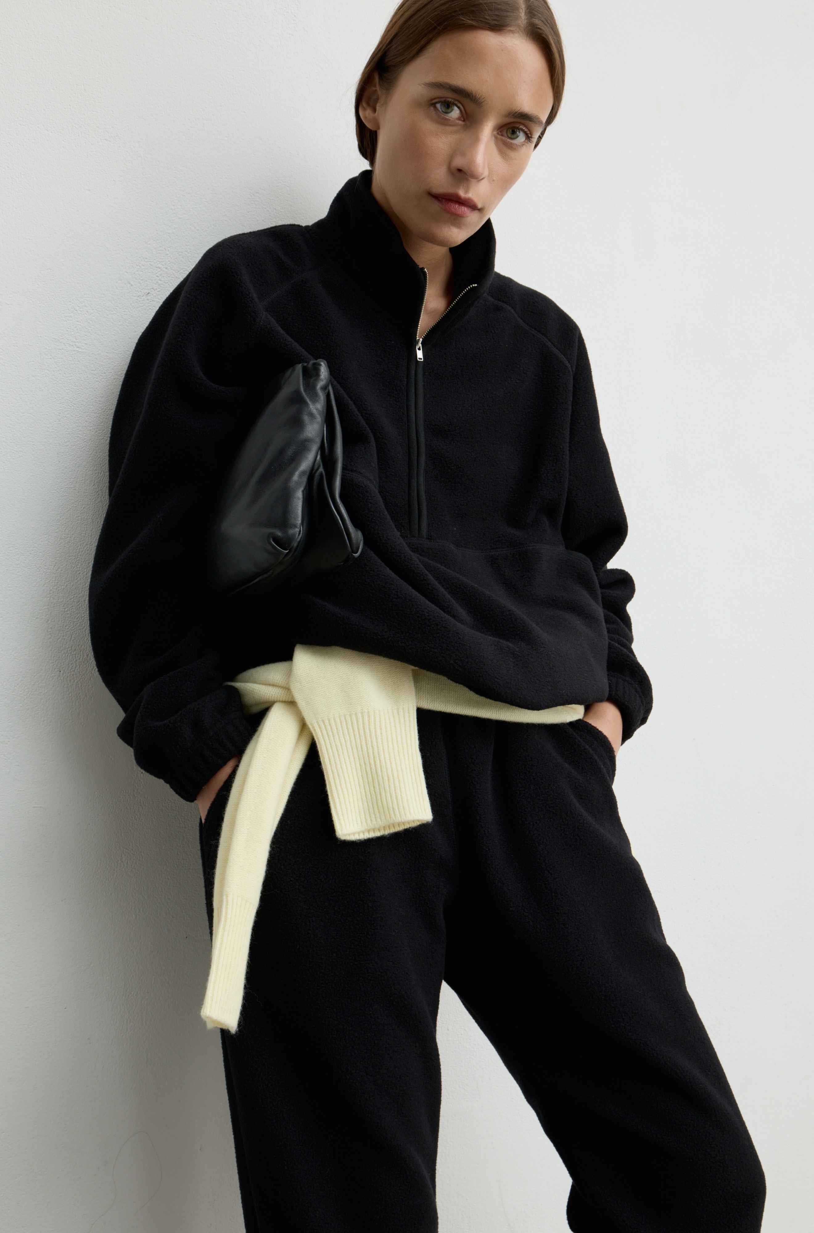 A model wearing a black fleece half-zip pullover, black sweatpants, and a cream ribbed knit belt tied at the waist, holding a black clutch against a light gray wall.