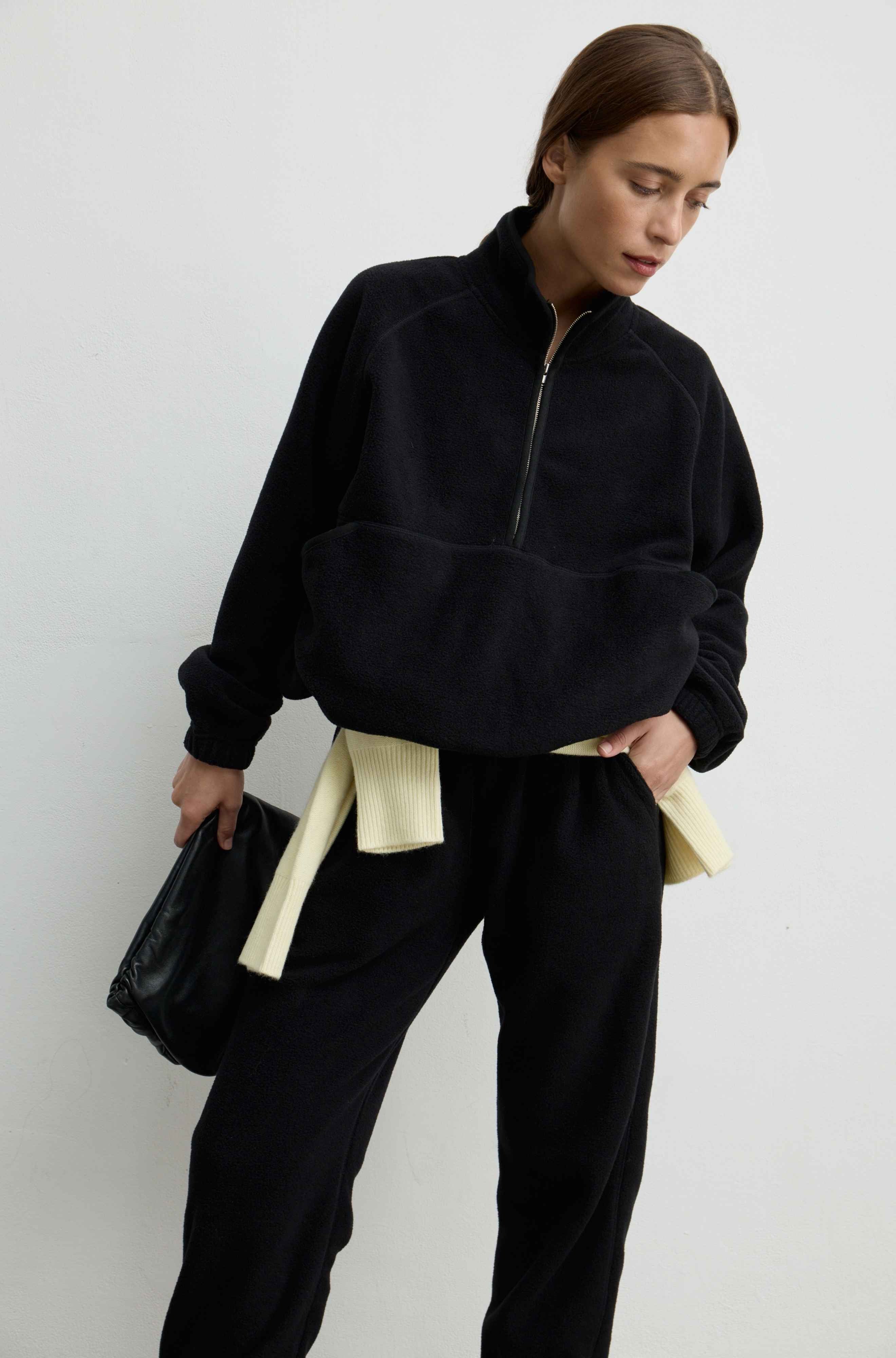 A woman modeling a black fuzzy quarter-zip fleece jacket with matching black sweatpants, featuring a cream-colored underlayer peeking out at the hem and waist, set against a plain light gray background.