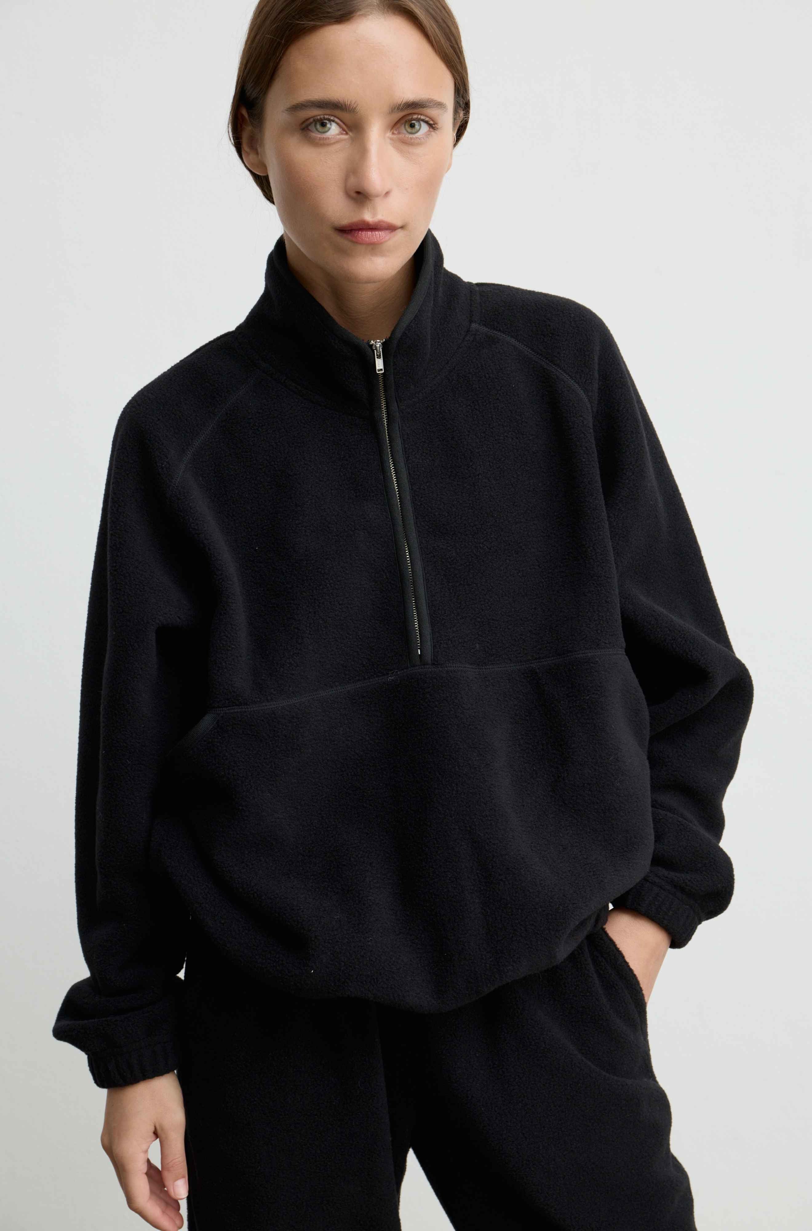 A woman wearing a black quarter-zip fleece pullover with a stand-up collar and front kangaroo pocket, paired with matching black pants, against a light gray studio background.