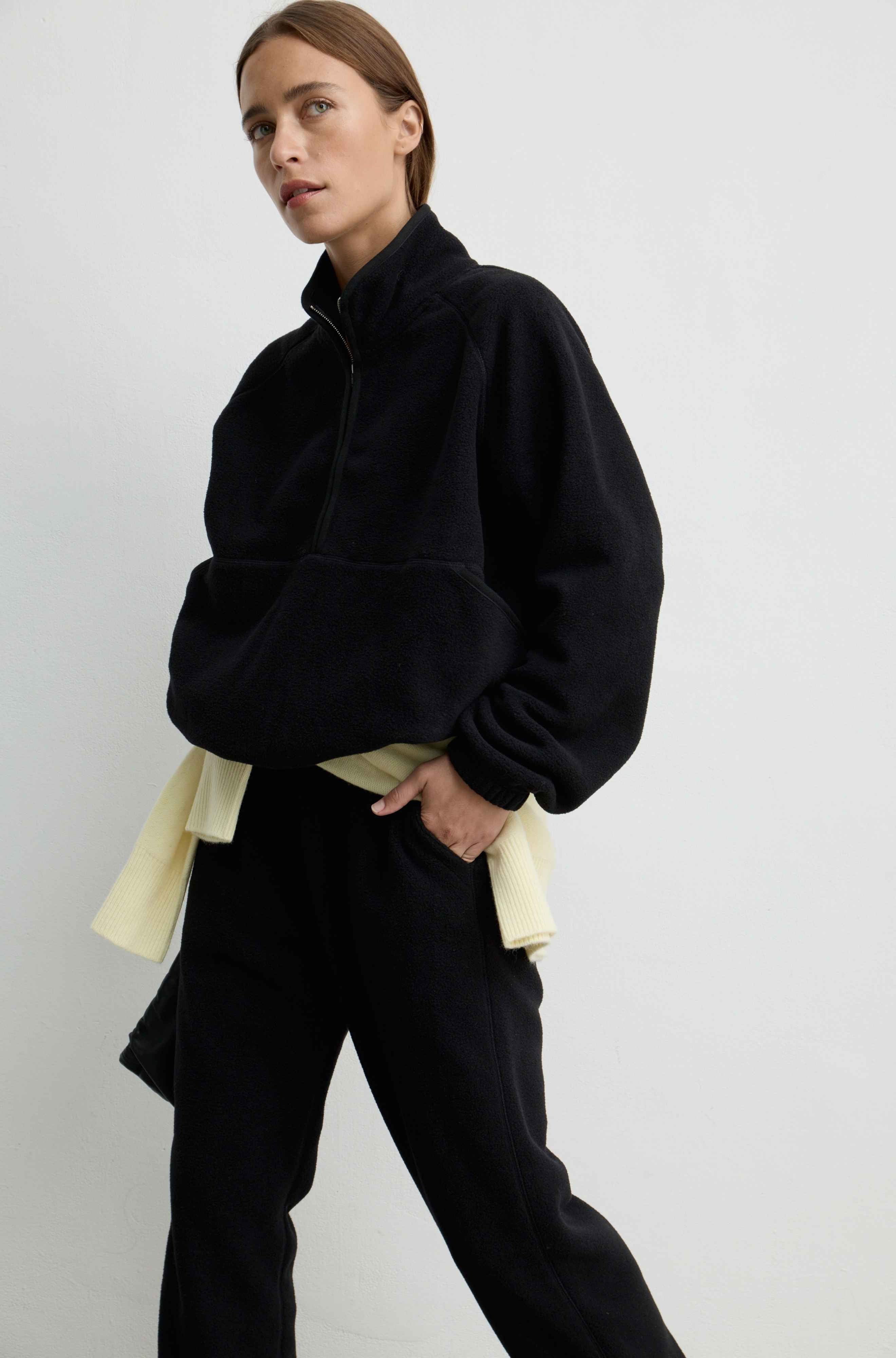 A model wearing a black half-zip fleece pullover with voluminous balloon sleeves, styled with black sweatpants and a cream shirt tied at the waist, against a plain light gray studio background.