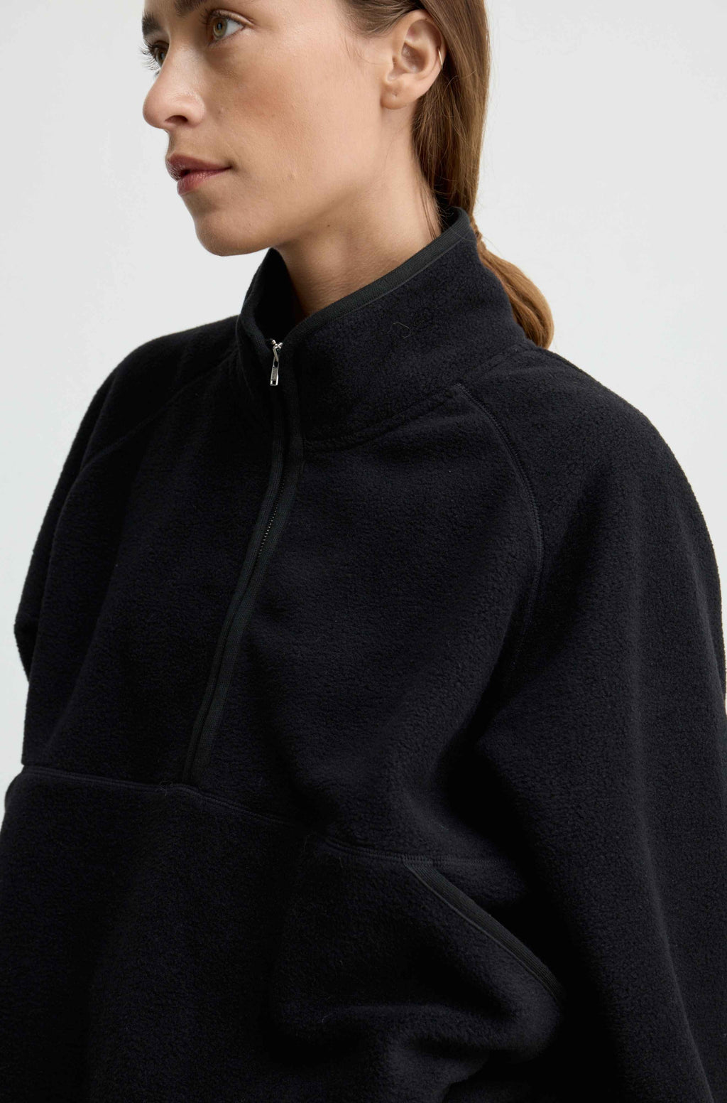 A close-up side profile of a woman wearing a dark navy fleece pullover jacket with a high collar and a half-zip front, set against a plain light gray background.