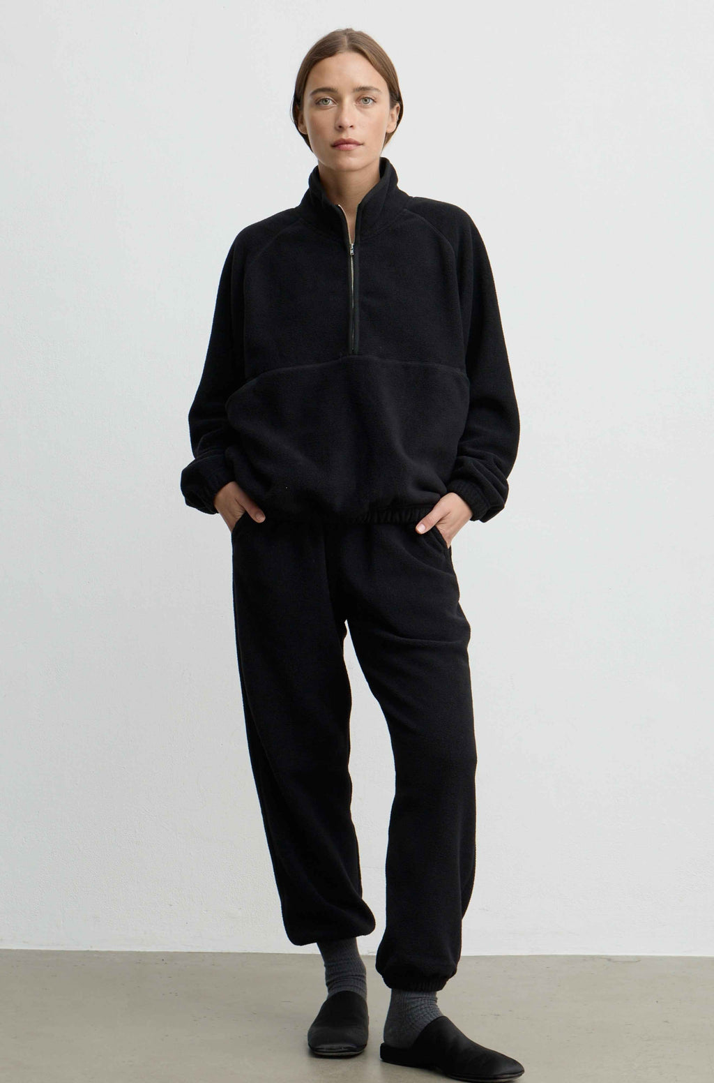 A woman modeling a black fleece tracksuit with a half-zip pullover top and matching jogger pants, standing against a plain light gray studio background.