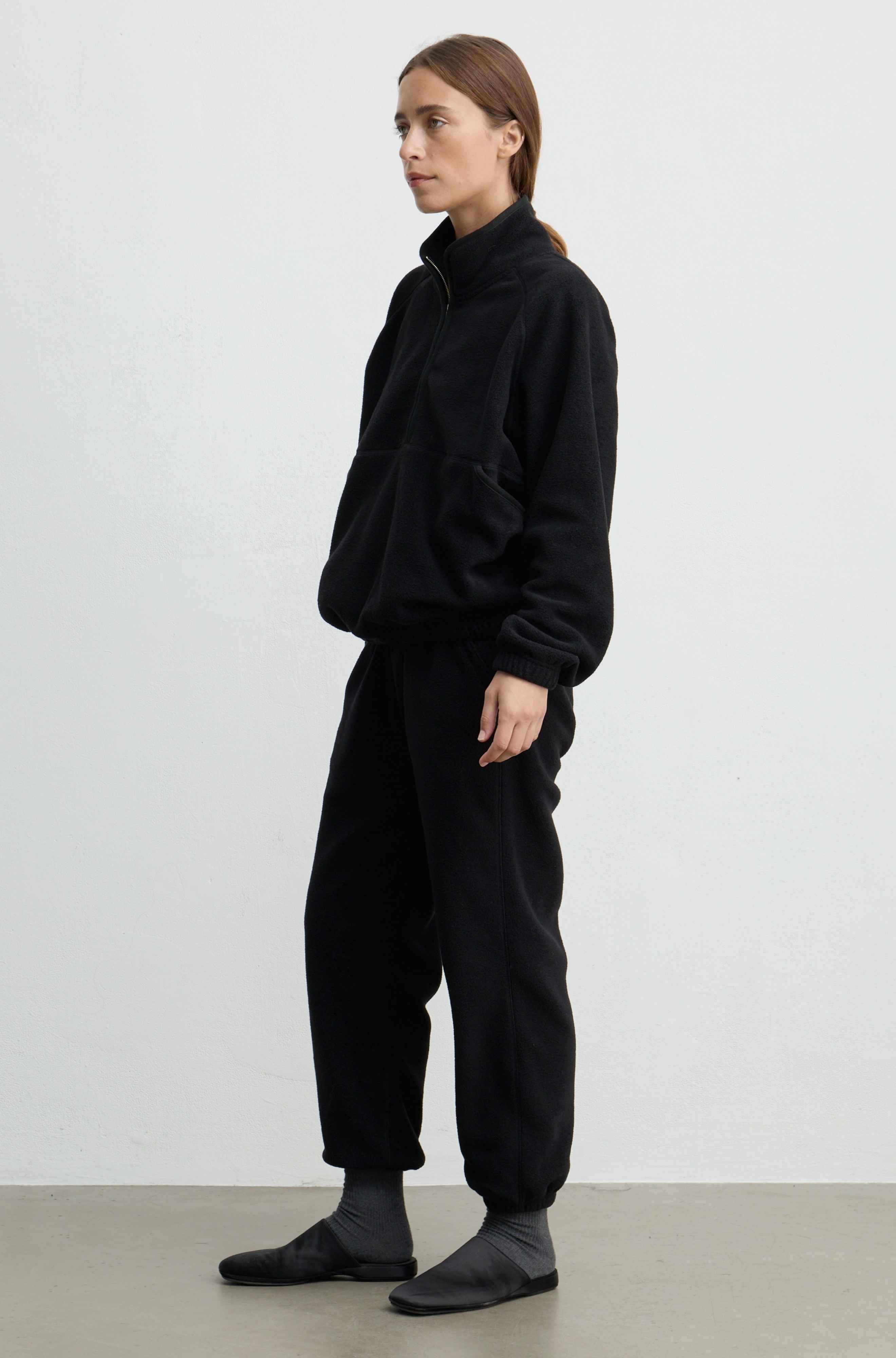 A woman modeling a black fleece zip-up jacket and matching black jogger pants against a plain light gray studio background.