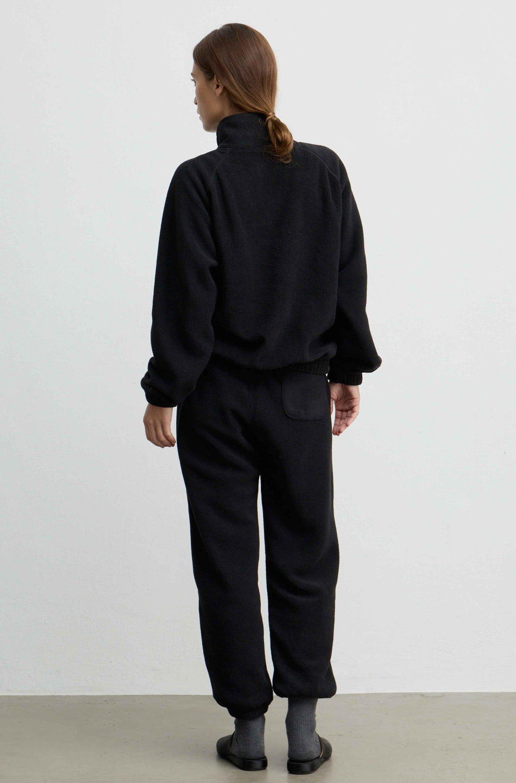 A model viewed from behind wearing a black fleece zip-up jacket and matching black fleece pants, standing against a plain white studio wall.