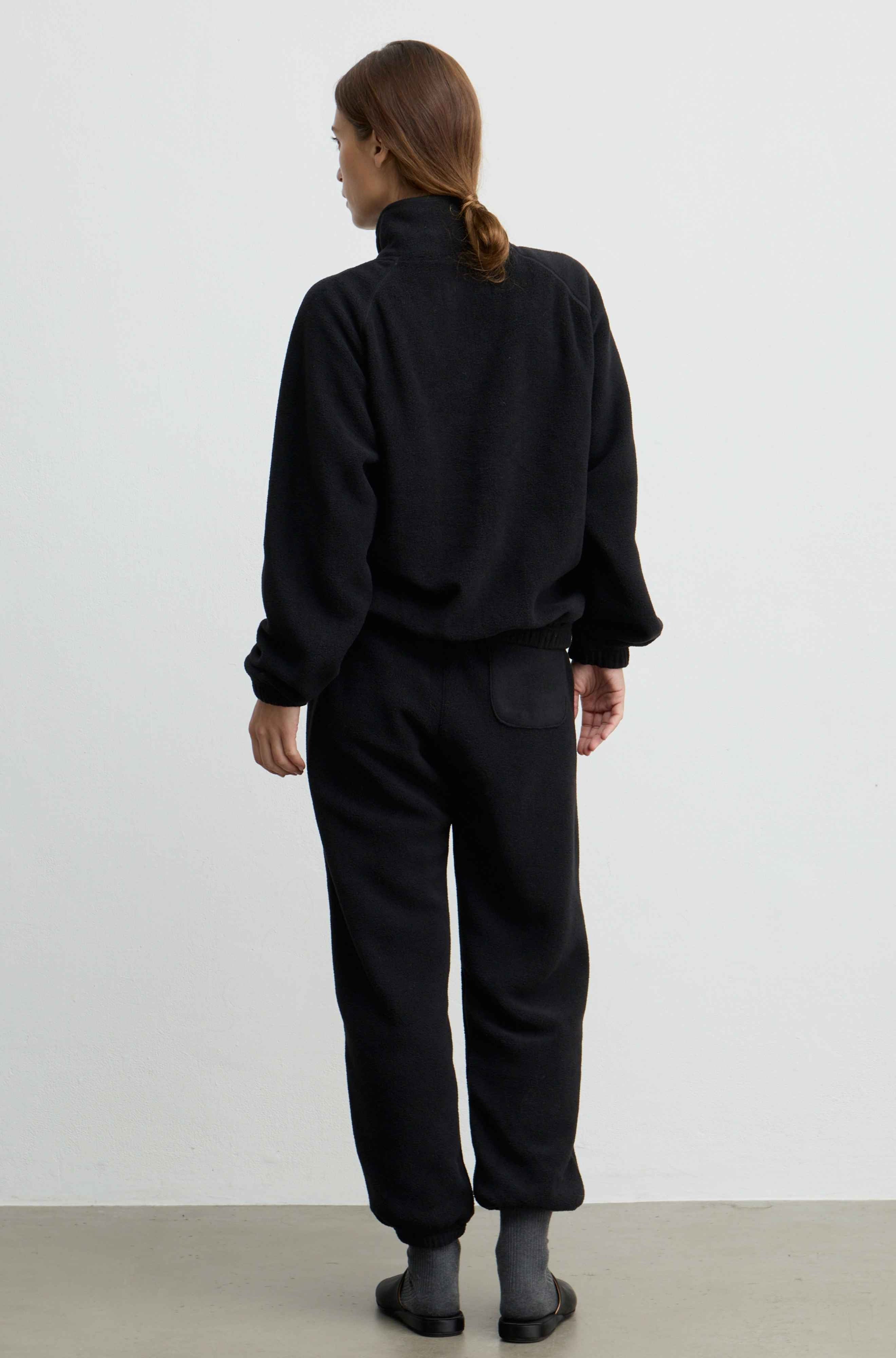 A model viewed from behind wearing a black fleece zip-up jacket and matching black fleece pants, standing against a plain white studio wall.