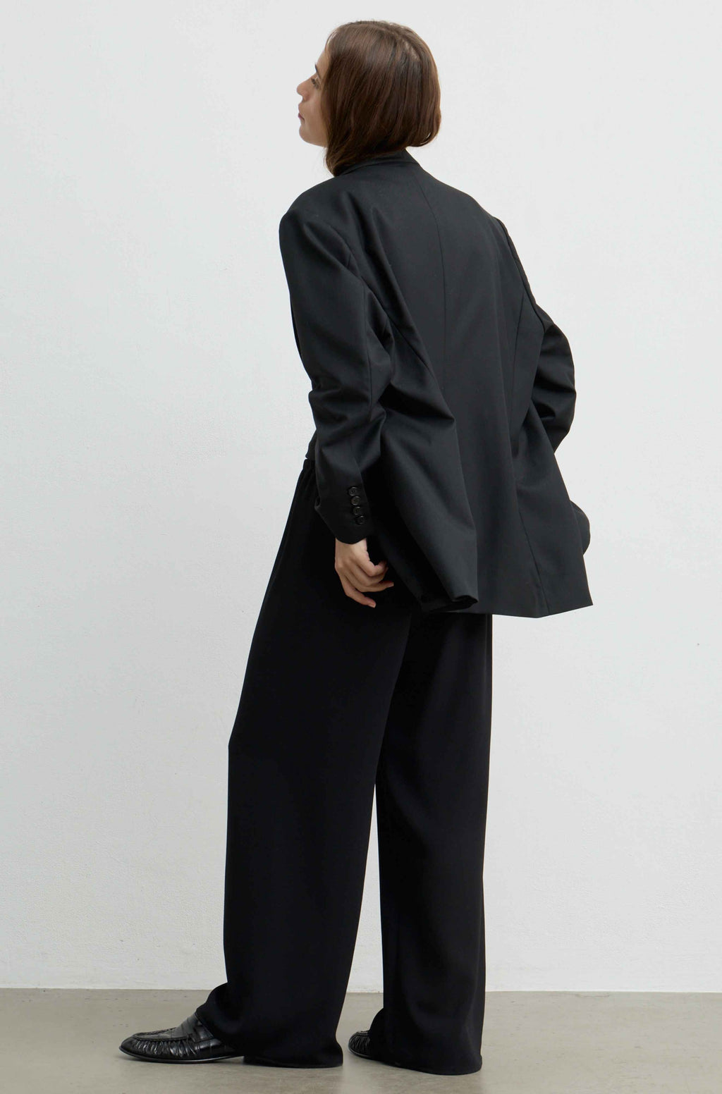 A model shown from the back wearing an oversized black blazer and matching wide-leg trousers, paired with black leather shoes, against a plain white studio background.