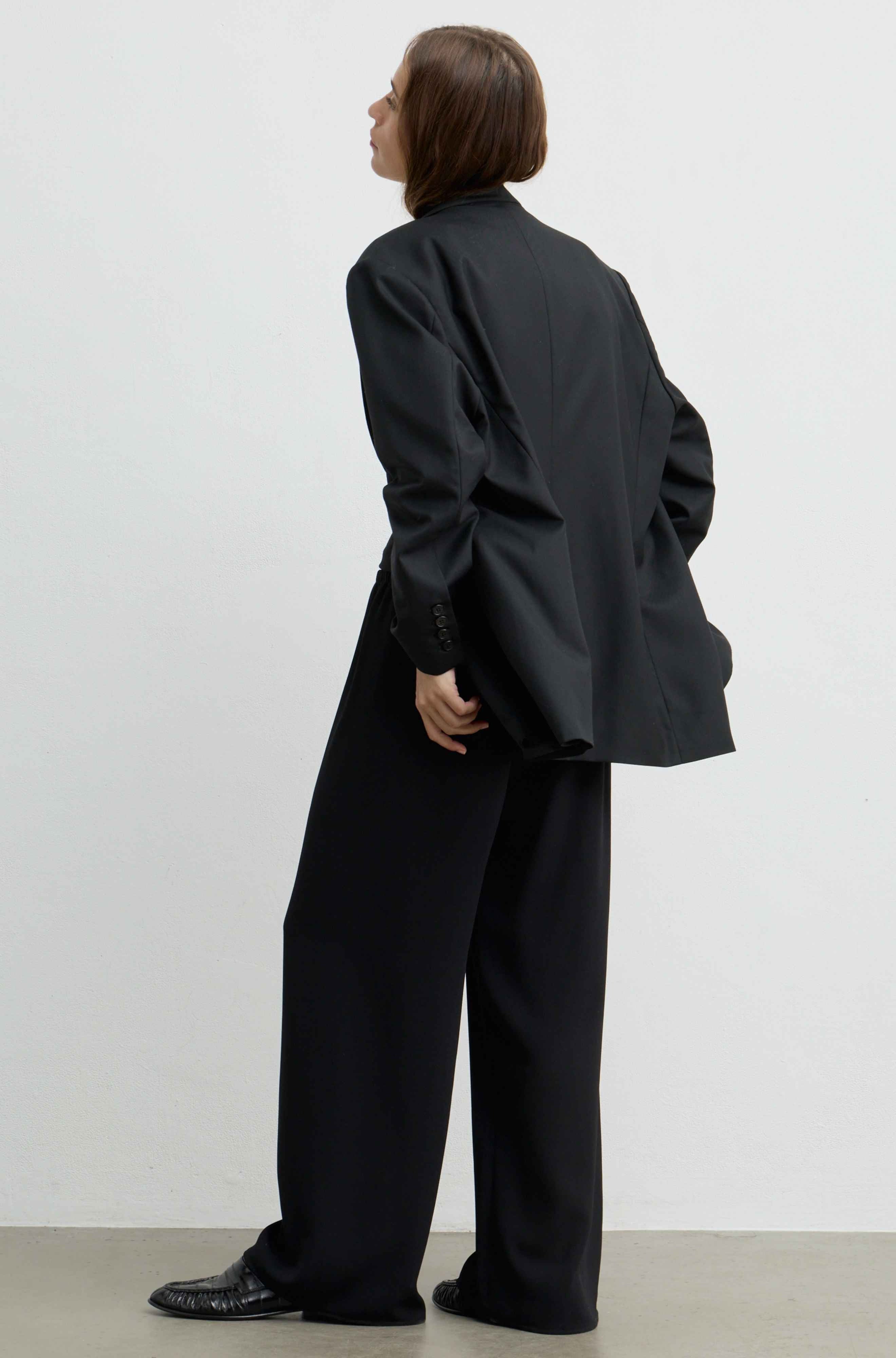 A model shown from the back wearing an oversized black blazer and matching wide-leg trousers, paired with black leather shoes, against a plain white studio background.
