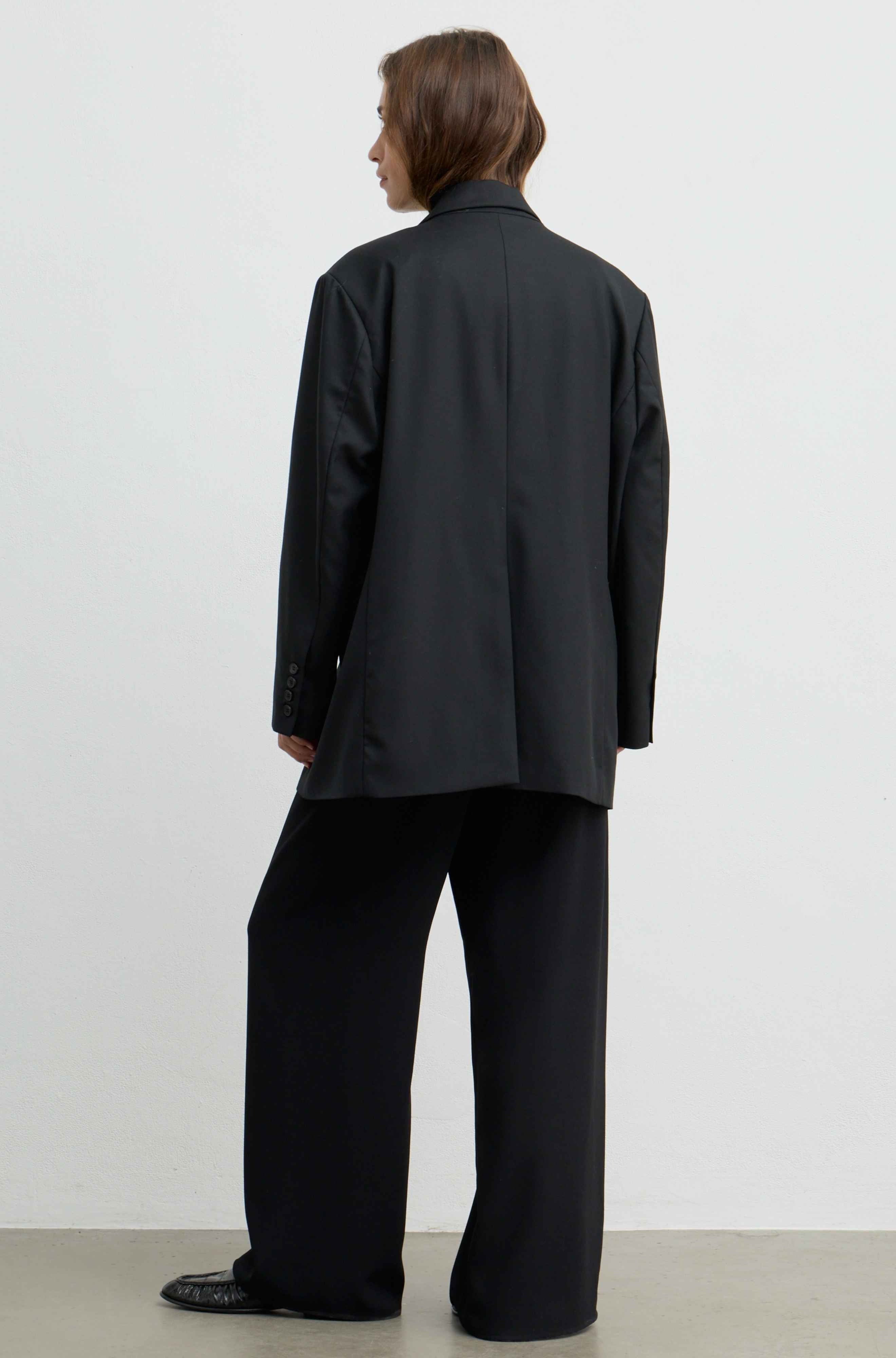 A model shown from the back wearing an oversized black blazer and matching wide-leg trousers against a plain white studio background.