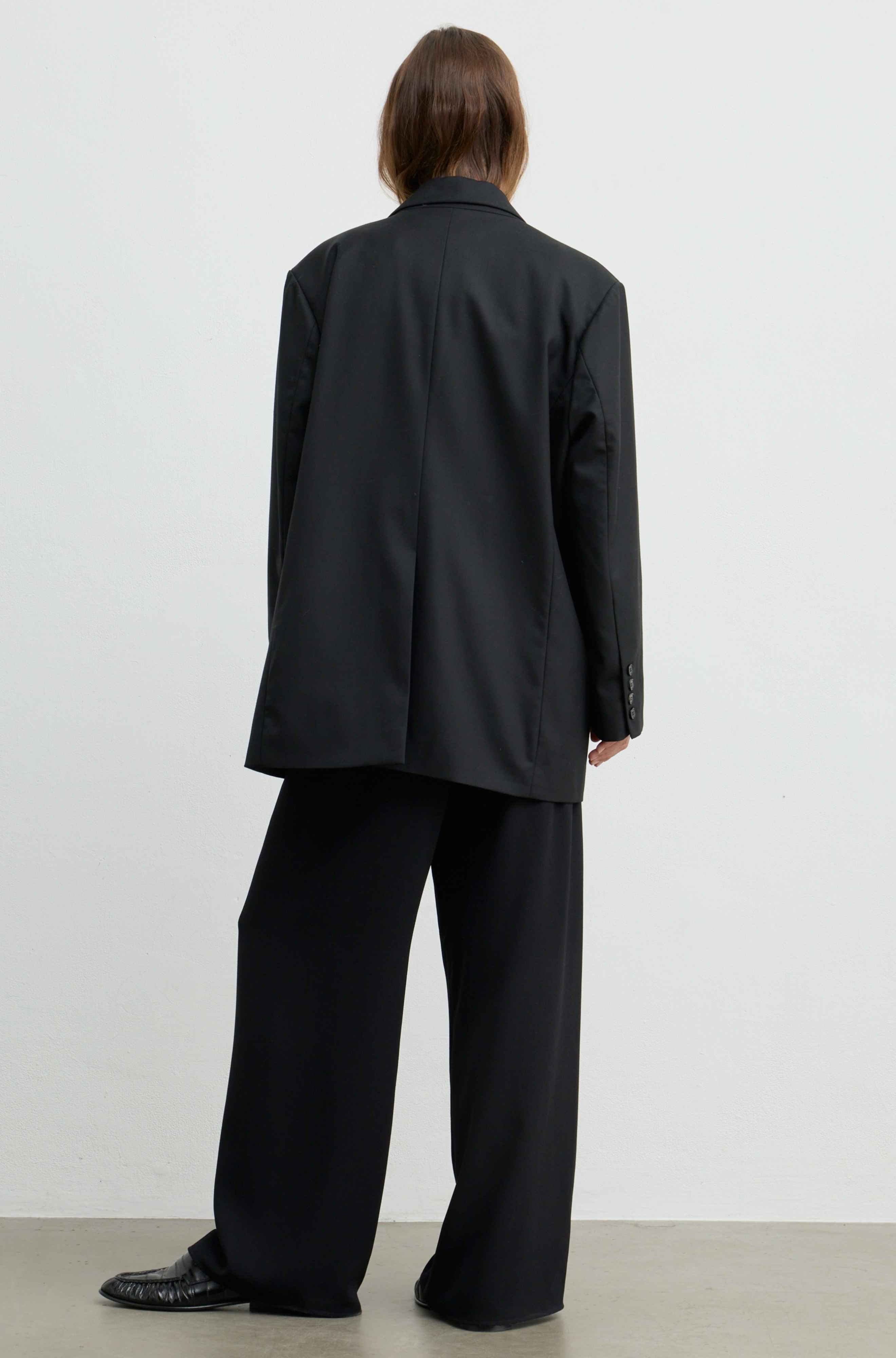 A model viewed from the back wearing an oversized black blazer and matching wide-leg trousers, standing against a plain light gray studio wall.