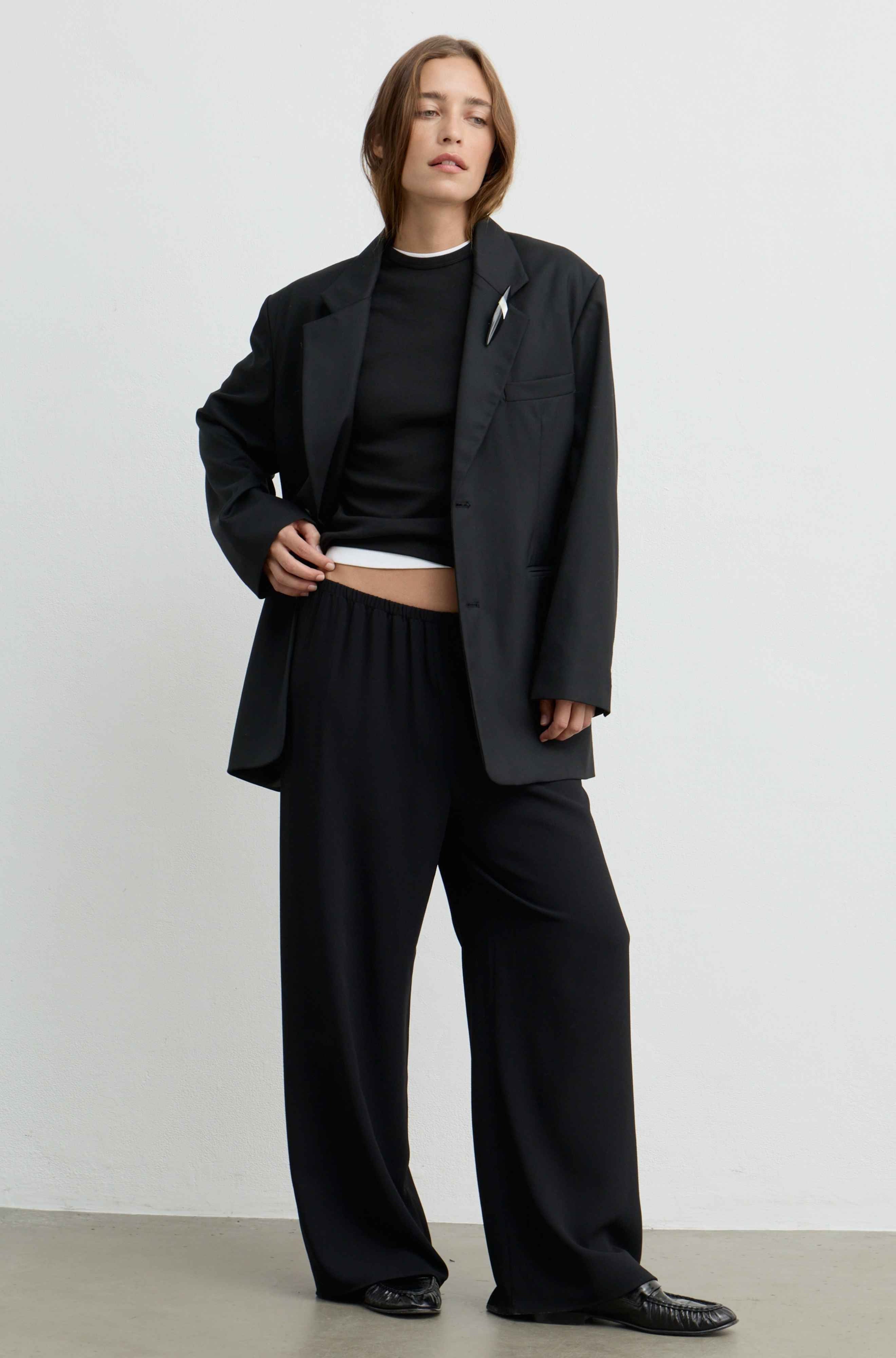 A model wearing a black oversized blazer, black wide-leg trousers, a black cropped top, and black leather loafers, standing against a plain light gray studio background.