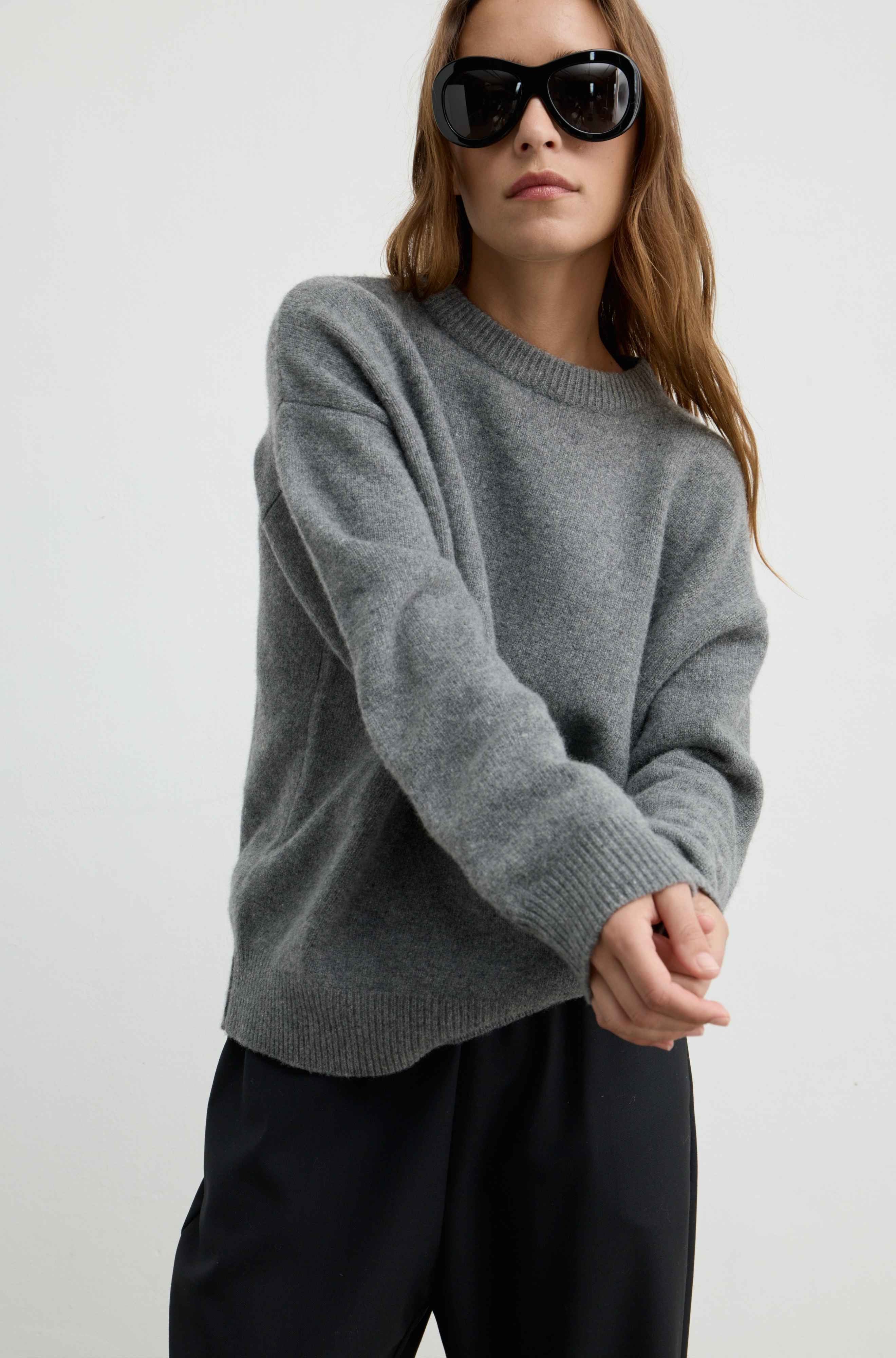 A woman wearing an oversized heather gray knit sweater and black trousers, styled with black sunglasses against a plain light gray studio background.