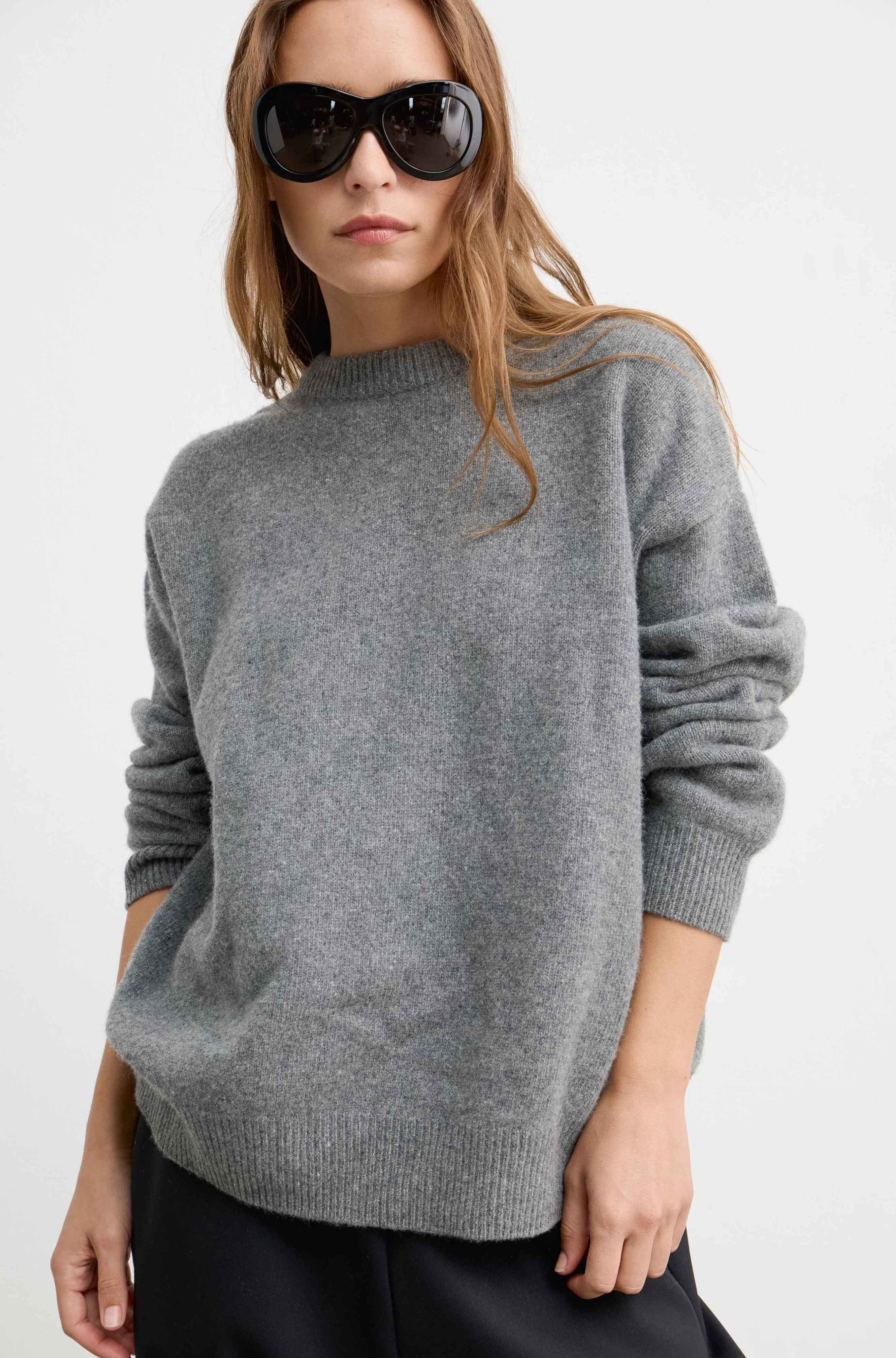 Woman wearing a wool and cashmere grey sweatshirt with oversized black sunglasses on a light grey background