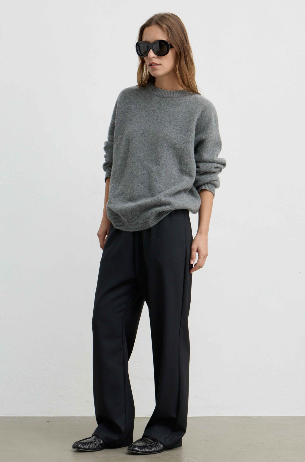 A woman modeling a gray knit crewneck sweater and black wide-leg trousers against a plain light gray studio background.