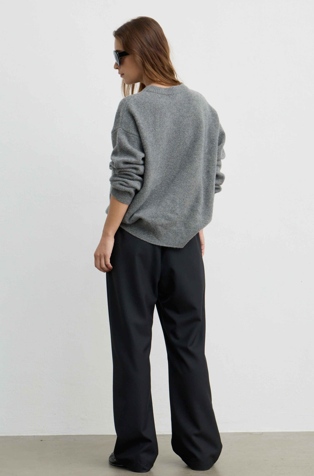 A woman shown from the back wearing a heather gray crewneck sweatshirt and black wide-leg trousers, standing against a plain white studio wall.