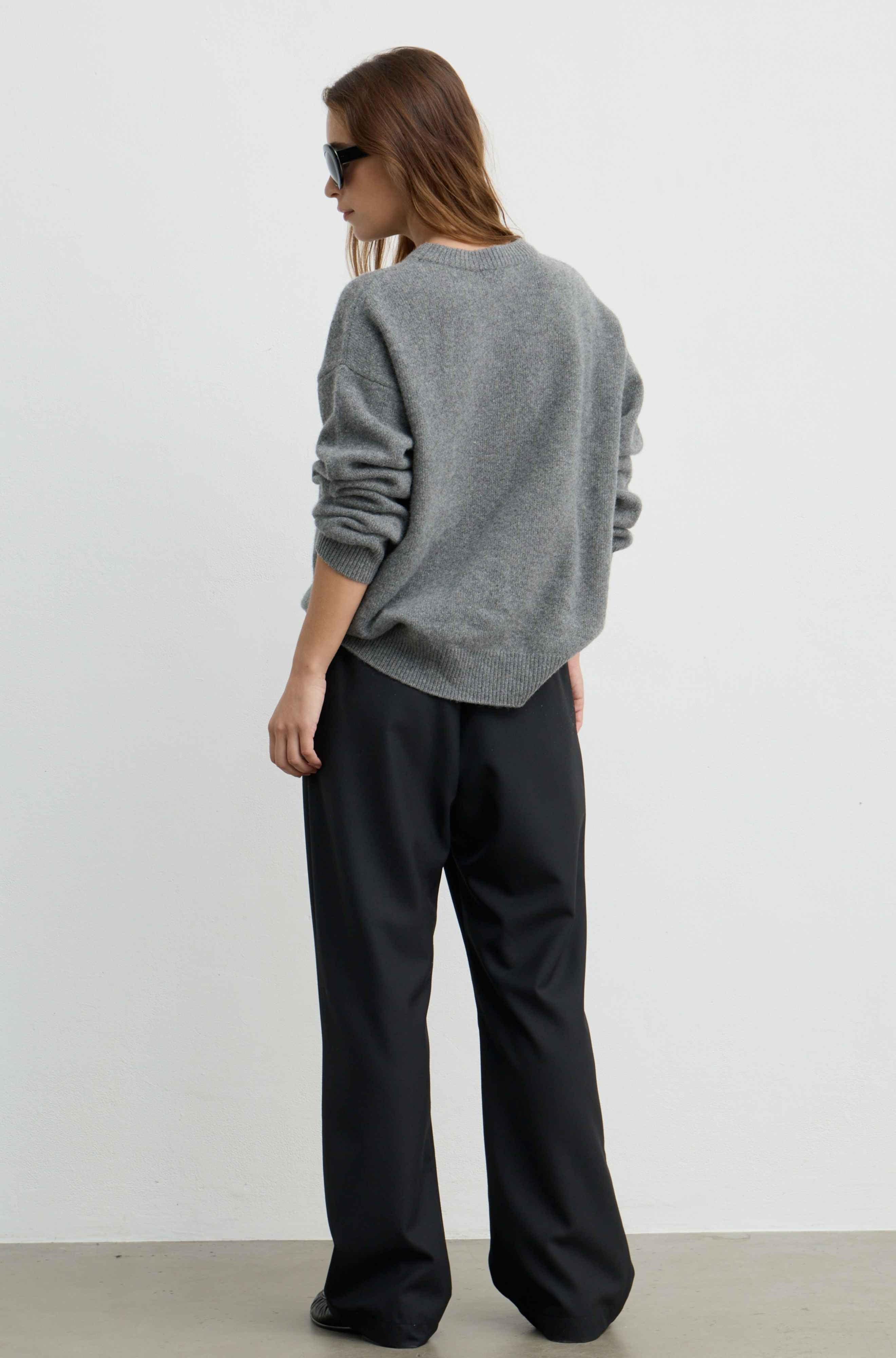 A woman shown from the back wearing a heather gray crewneck sweatshirt and black wide-leg trousers, standing against a plain white studio wall.