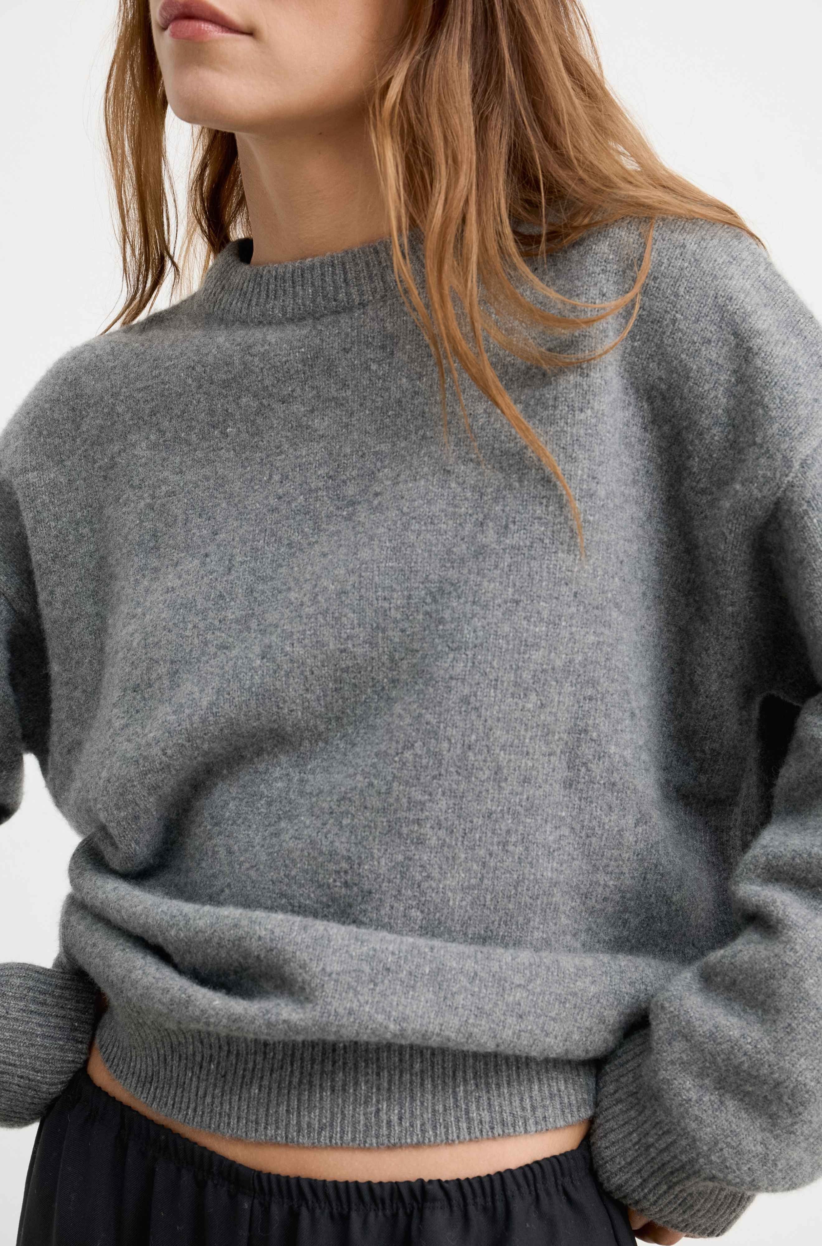 A close-up of a woman wearing a heather gray knit crewneck sweater, showing the texture of the fabric and the relaxed fit of the garment.