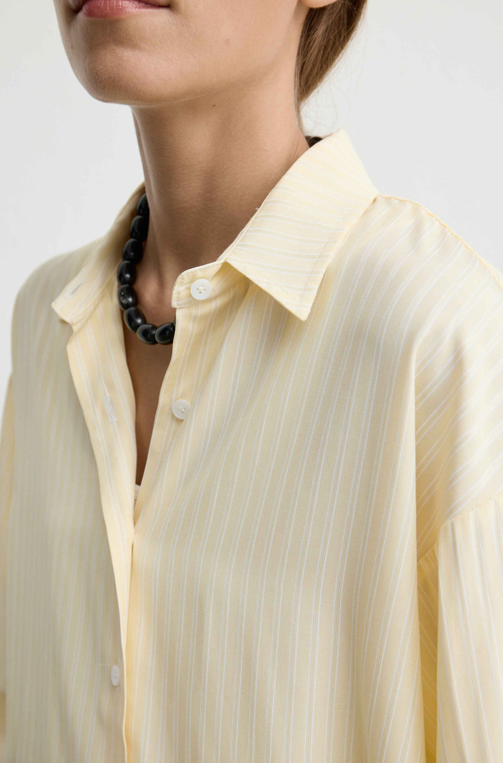 A close-up of a model wearing a pale yellow button-up shirt with thin vertical white stripes, styled with a black beaded necklace against a light gray background.
