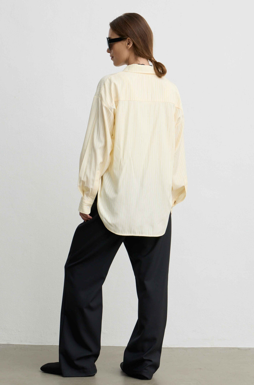 A woman shown from the back wearing an oversized cream-colored button-up shirt and loose black trousers, standing against a plain light gray wall.