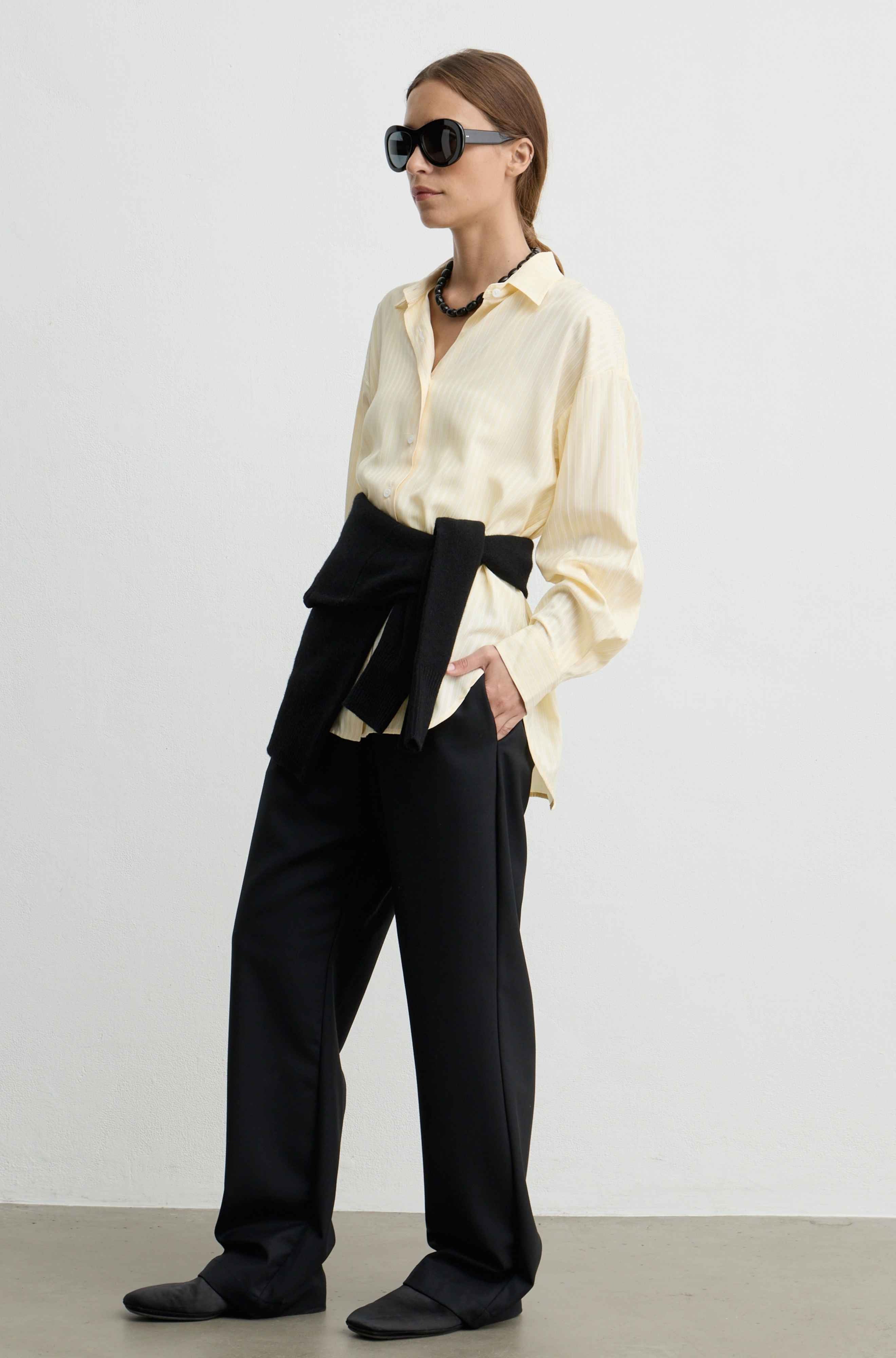 A model wearing a pale yellow button-down shirt, black wide-leg trousers with a large black waist tie, black sunglasses, and black slip-on shoes, standing against a plain light gray studio background.
