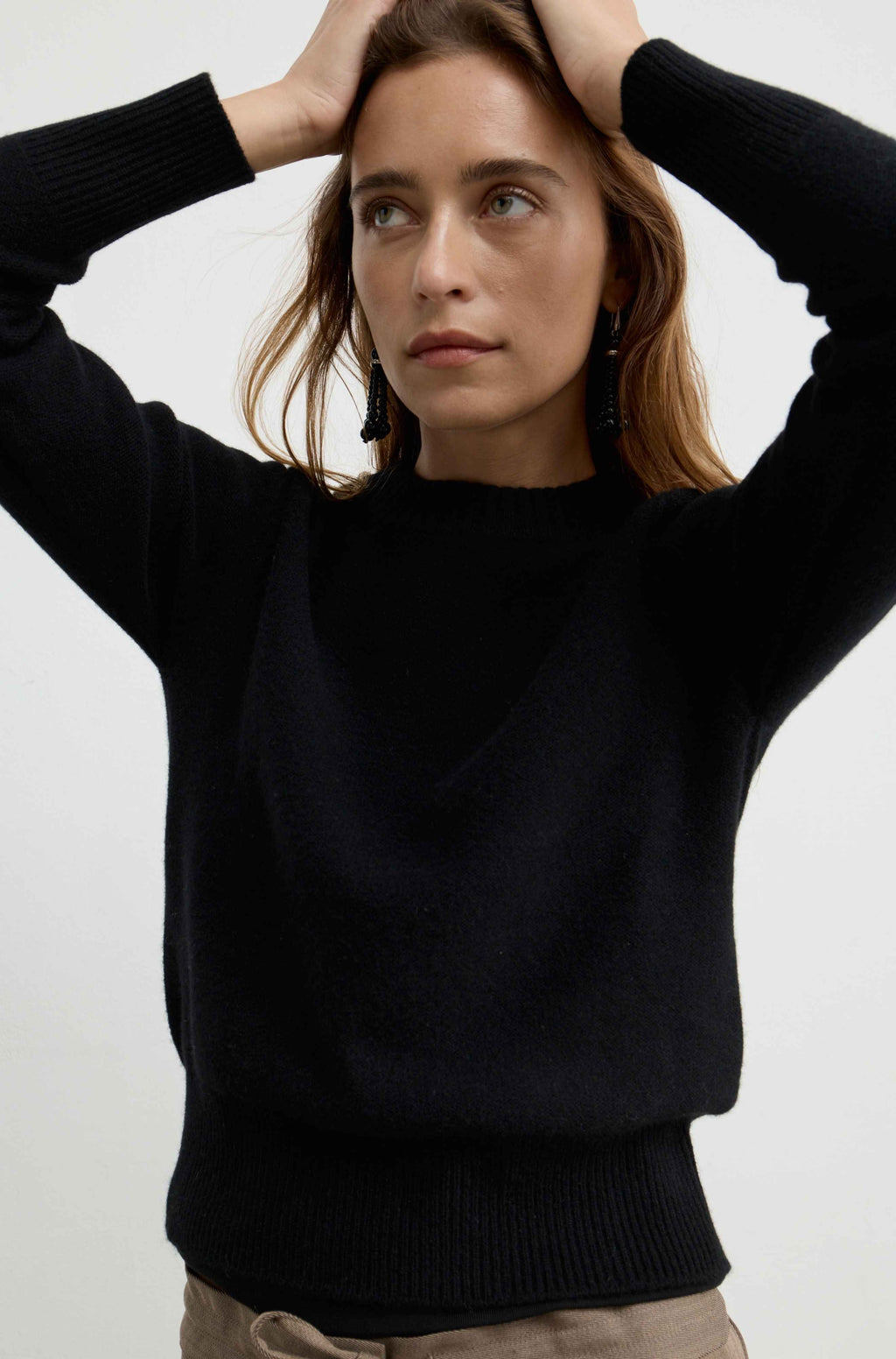 A woman wearing a black long-sleeve knit sweater, posing with her hands on her head against a plain light gray background.