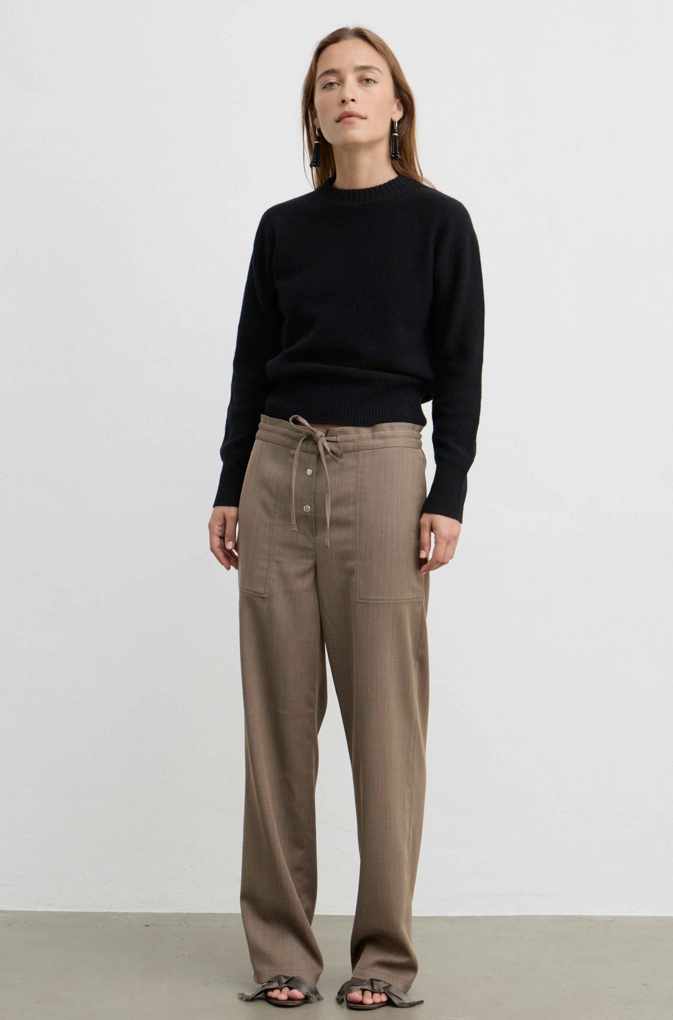 A woman stands against a plain light gray studio wall wearing a black long-sleeve knit sweater and high-waisted taupe-brown wide-leg trousers with a drawstring waist and front buttons. She is wearing dark sandals.