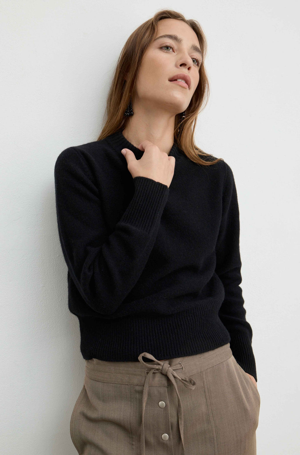 A woman wearing a black ribbed knit sweater and beige drawstring trousers, posing against a plain white wall.
