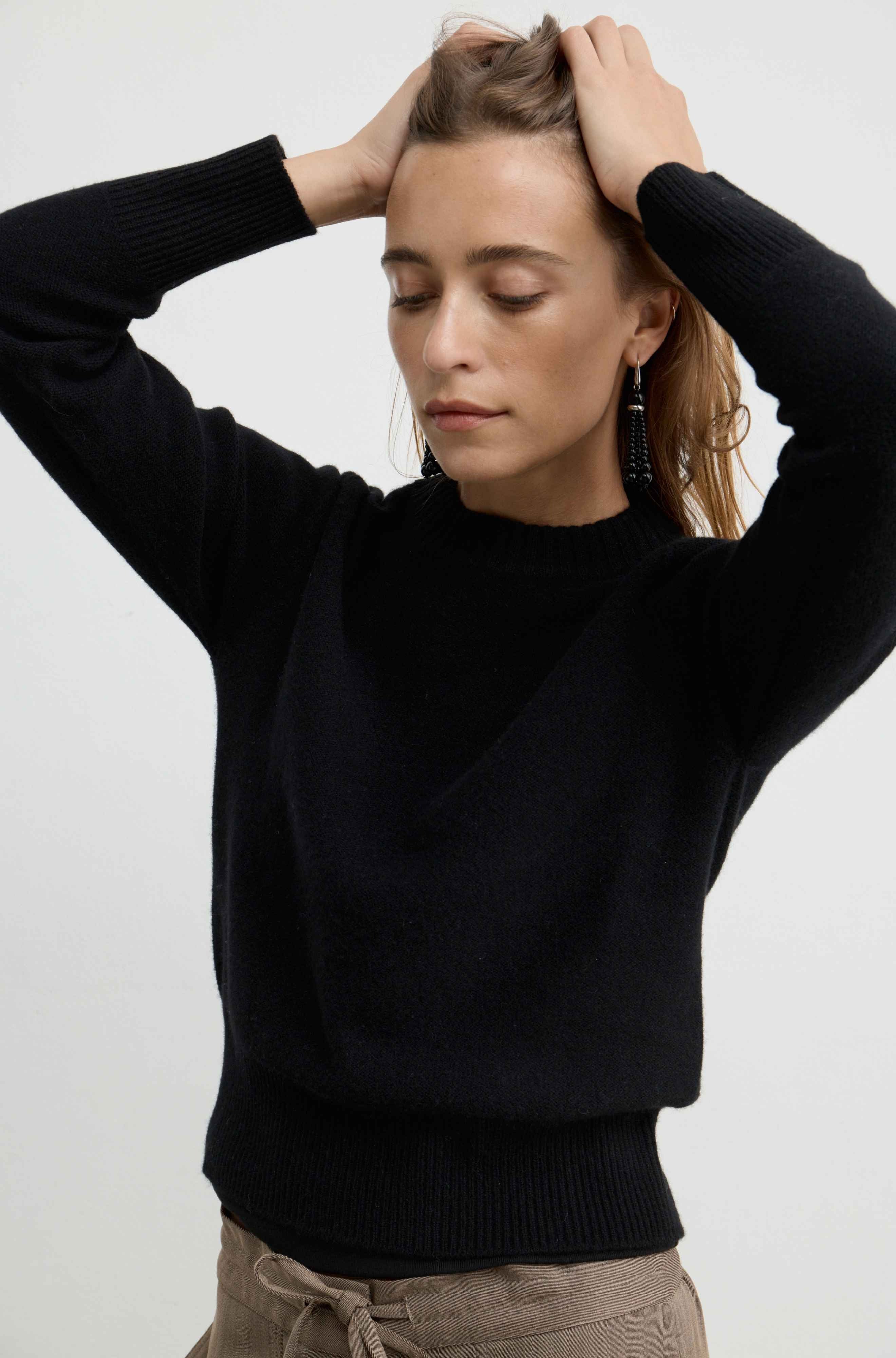 A woman wearing a black long-sleeve knit sweater and beige drawstring pants, posing with her hands in her hair against a plain light gray background.