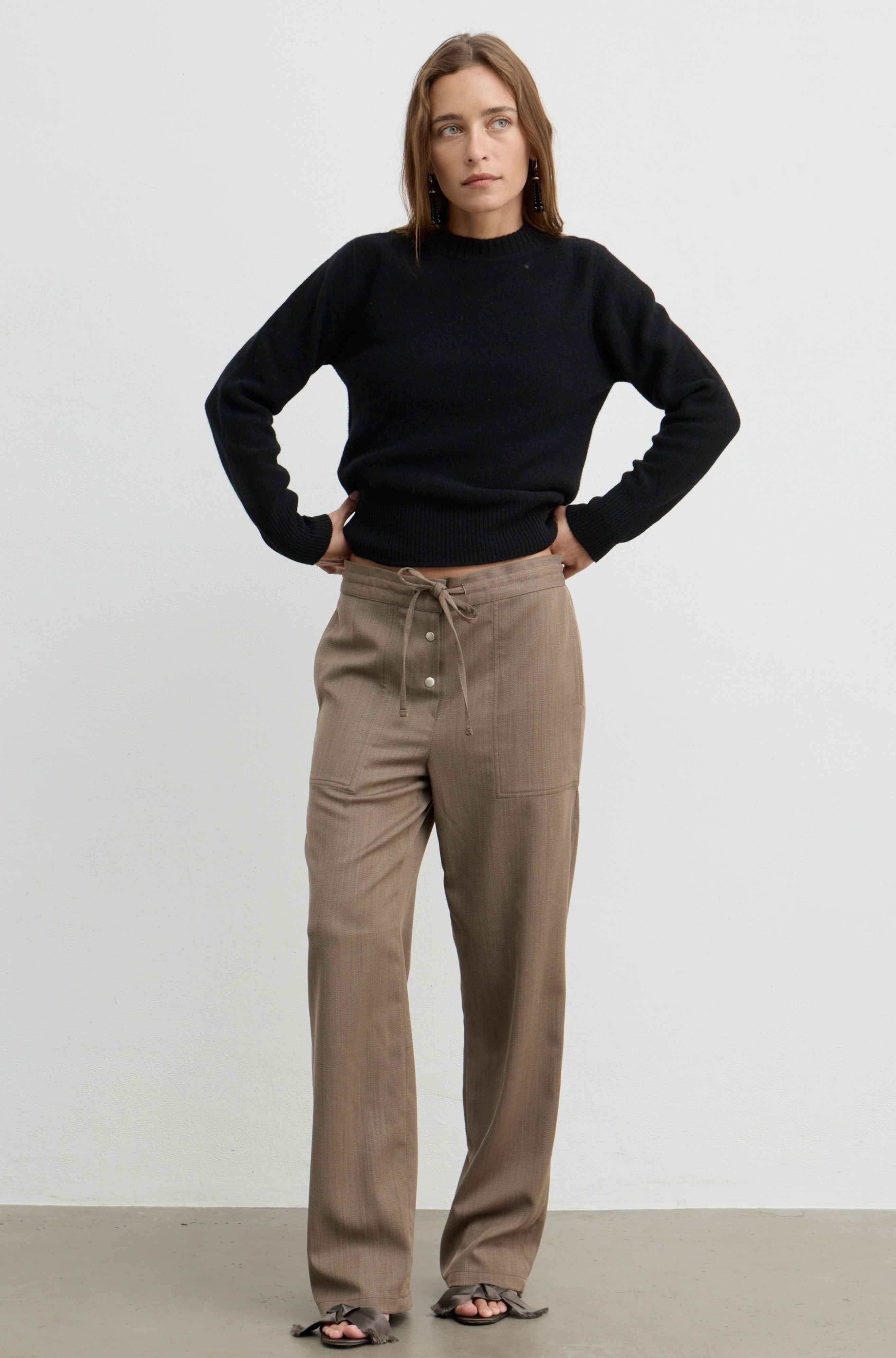 A woman modeling a black long-sleeve mock neck sweater and high-waisted taupe drawstring trousers against a plain white studio background.