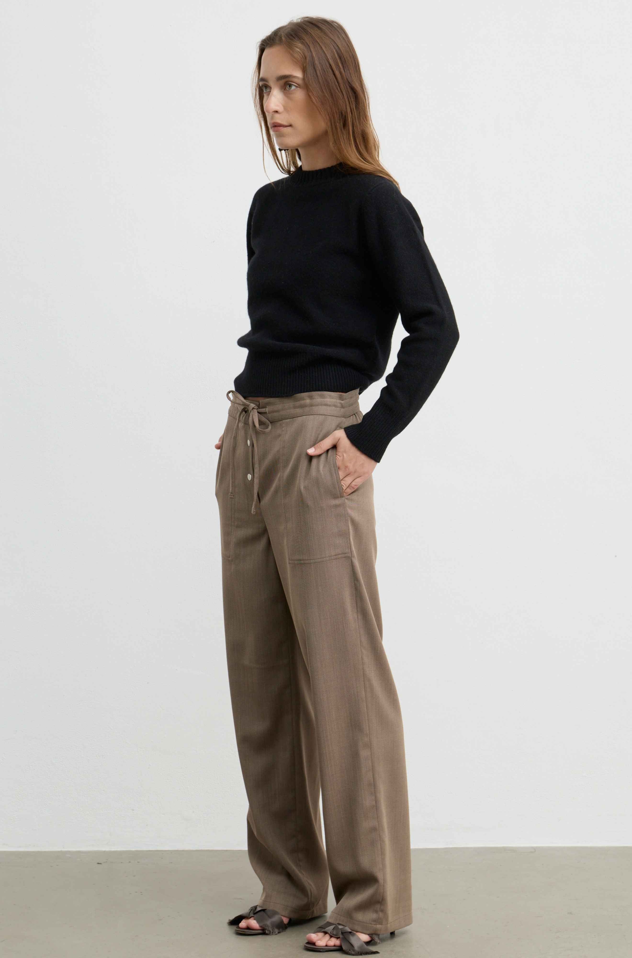 A woman modeling a black mock-neck long-sleeve sweater and high-waisted, wide-leg taupe-brown drawstring trousers, paired with black sandals, against a plain white studio background.