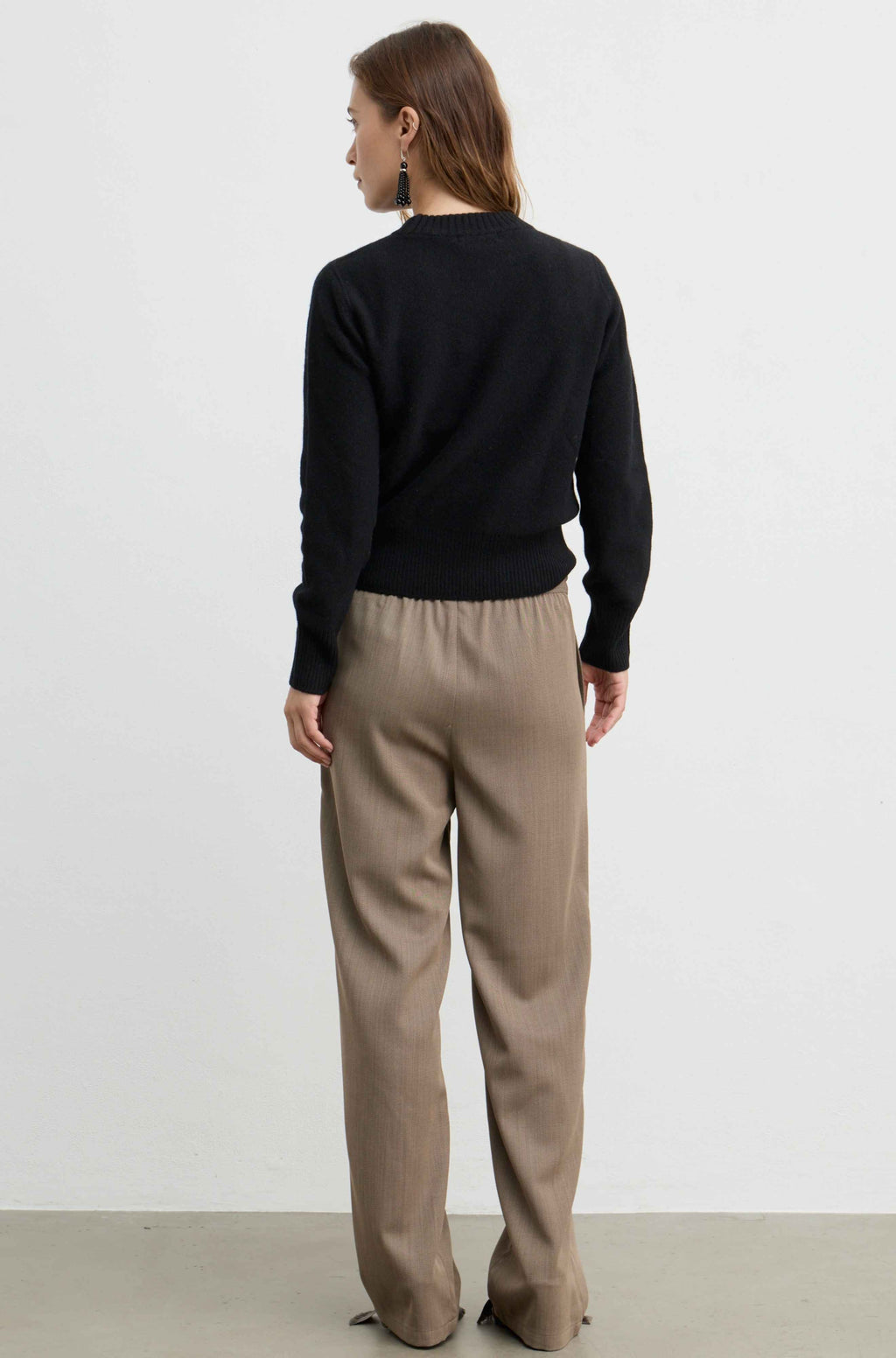 A woman is shown from the back wearing a black long-sleeve knit sweater and beige wide-leg trousers against a plain white studio background.