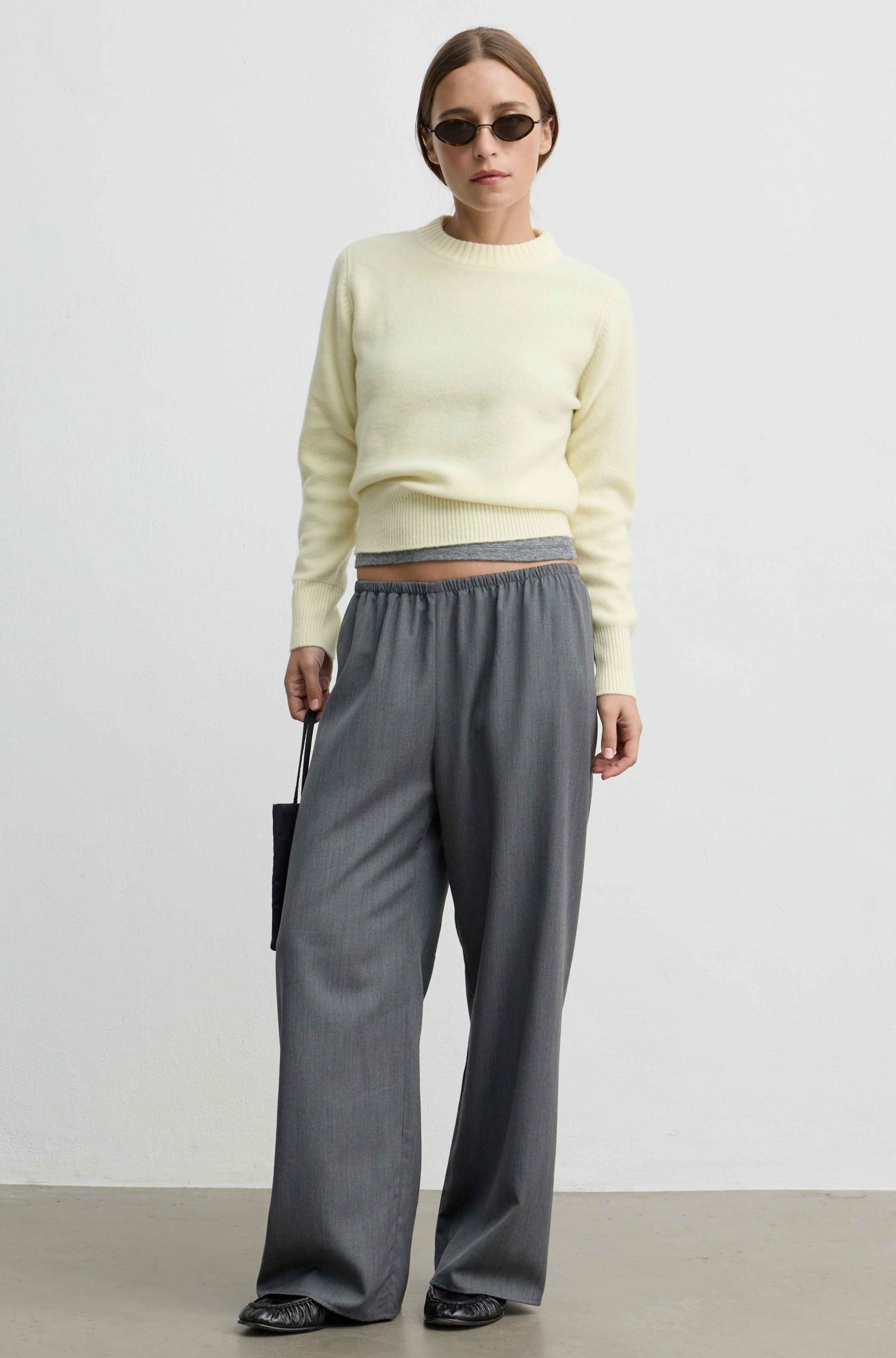 A model stands against a plain light gray studio wall wearing a pale yellow knit sweater, loose gray wide-leg trousers, black pointed shoes, and small dark sunglasses, while holding a small black handbag.