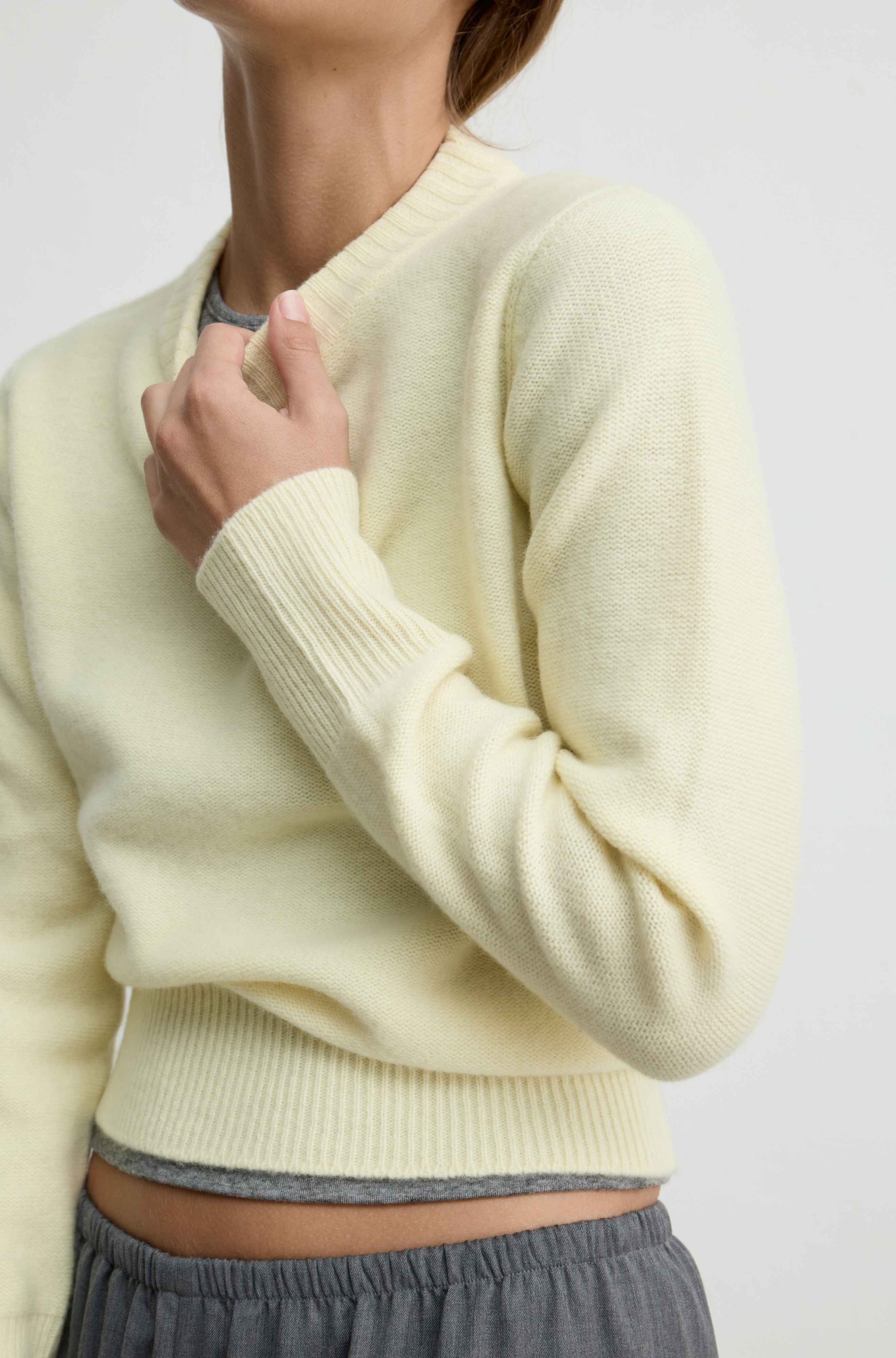 A close-up of a person wearing a pale yellow knit sweater with ribbed collar, cuffs, and hem, paired with gray drawstring pants against a light gray studio background.