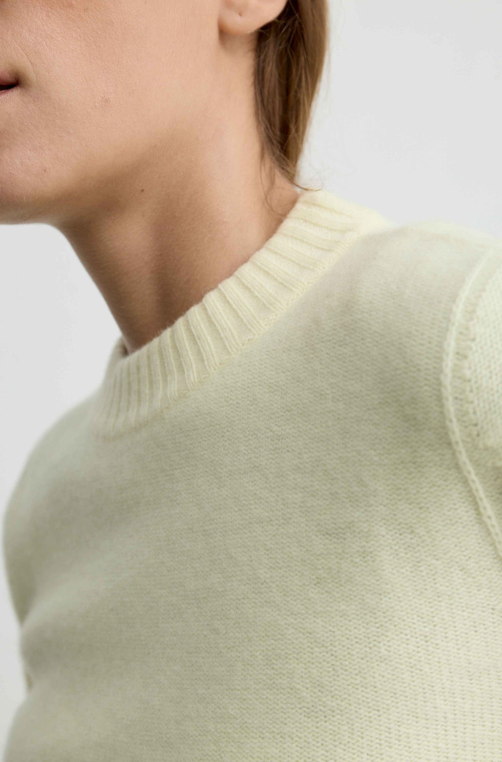 Close-up of a woman wearing a pale yellow knit sweater with a ribbed crew neck, shown against a light neutral background.