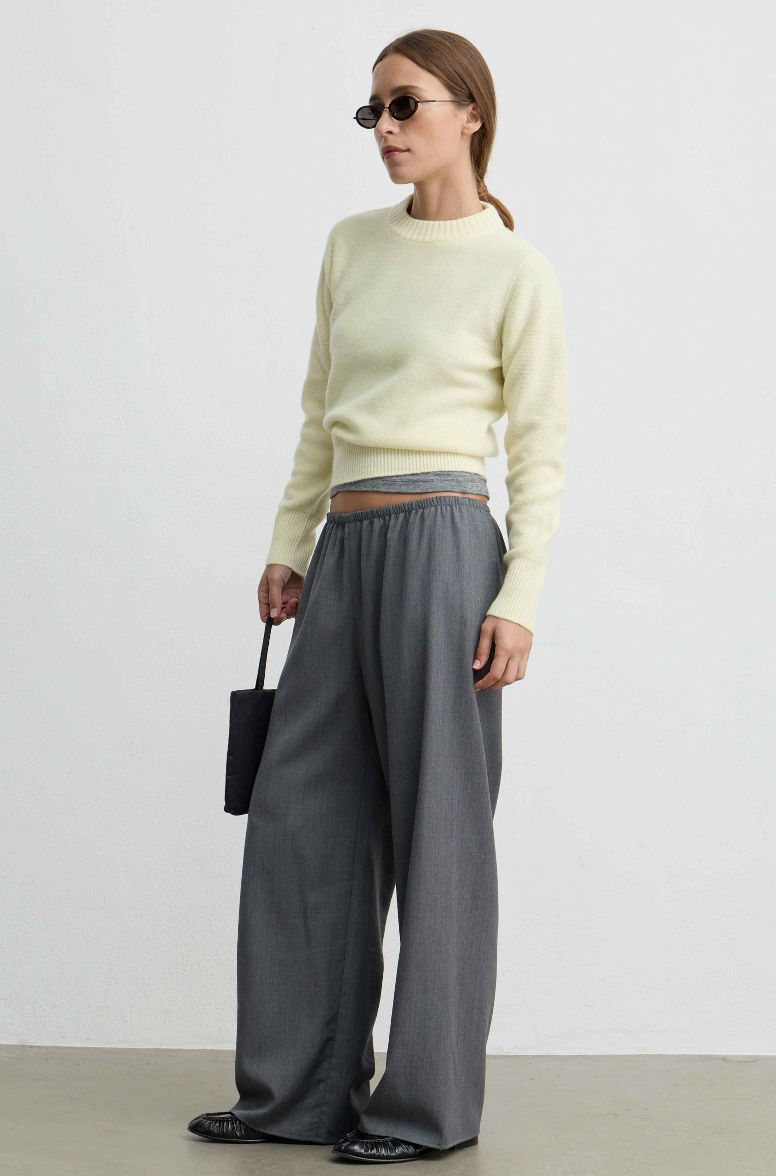 A woman modeling a pale yellow knit sweater and wide-leg gray trousers, standing against a plain light gray studio wall and floor. She is wearing dark sunglasses and holding a small black handbag.