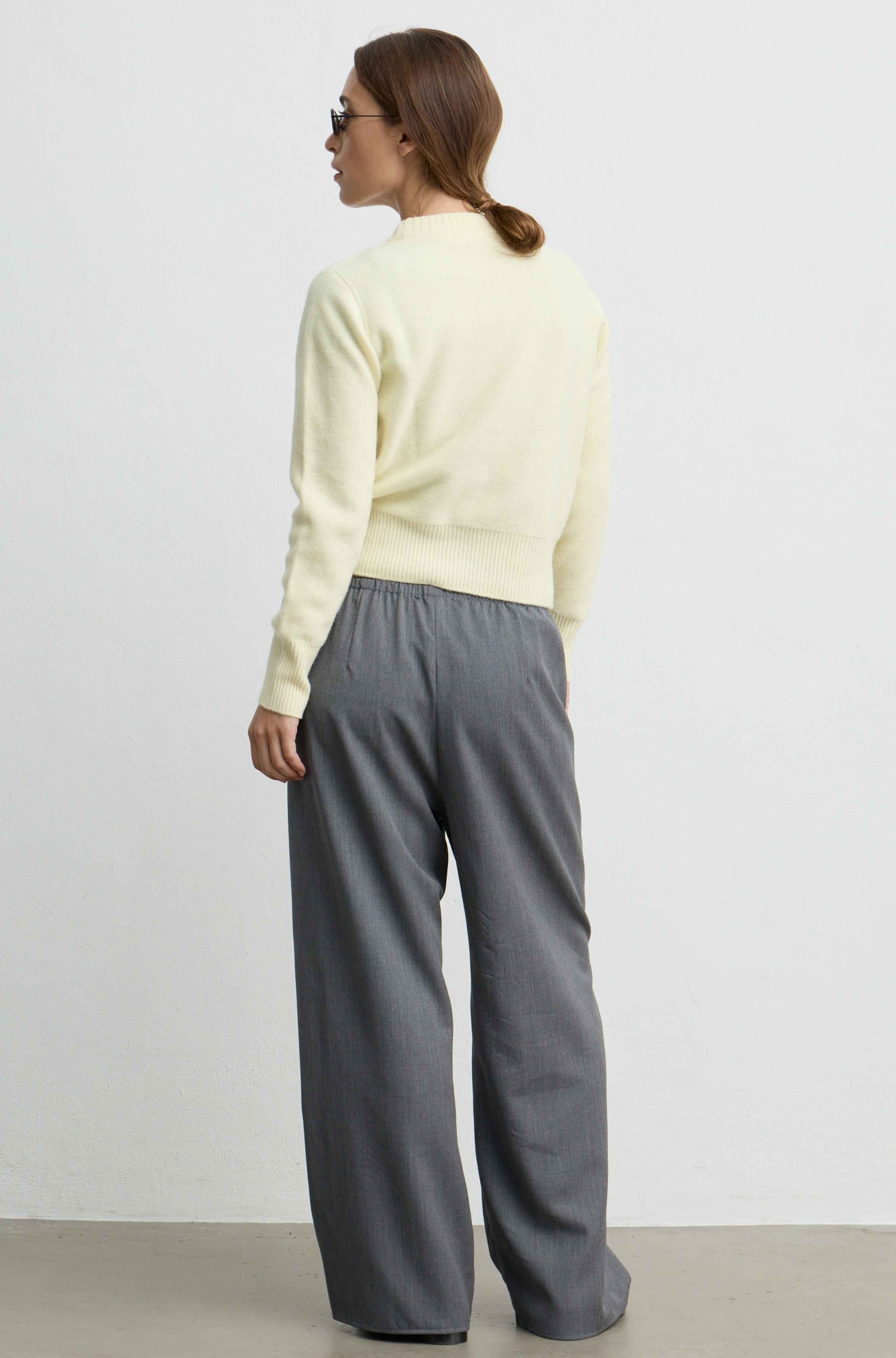A woman shown from the back wearing a pale yellow knit sweater and wide-leg gray trousers, standing against a plain light gray studio wall.