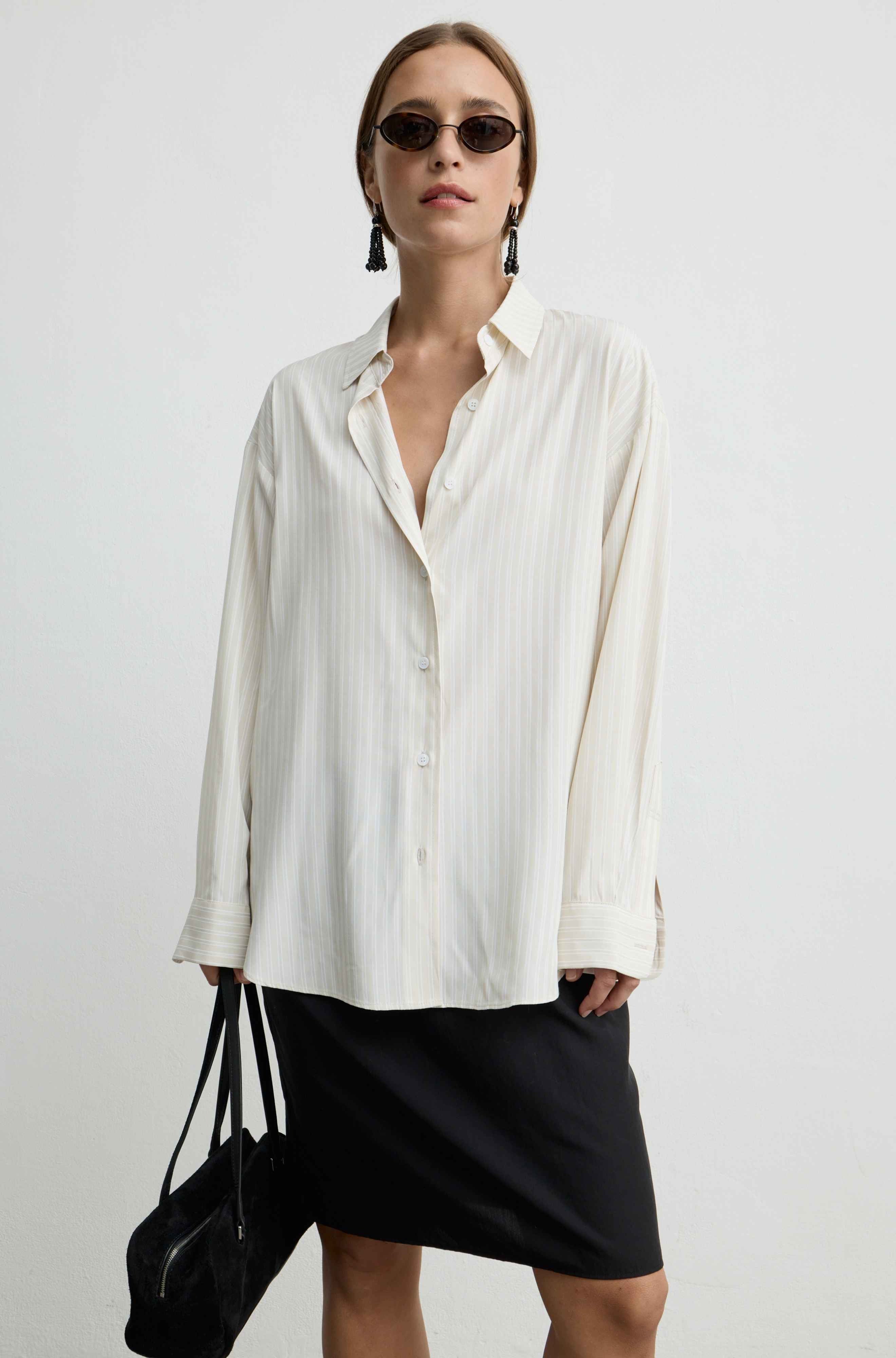 A woman modeling an oversized off-white striped button-up shirt with a black mini skirt and sunglasses, holding a black handbag against a plain light gray background.