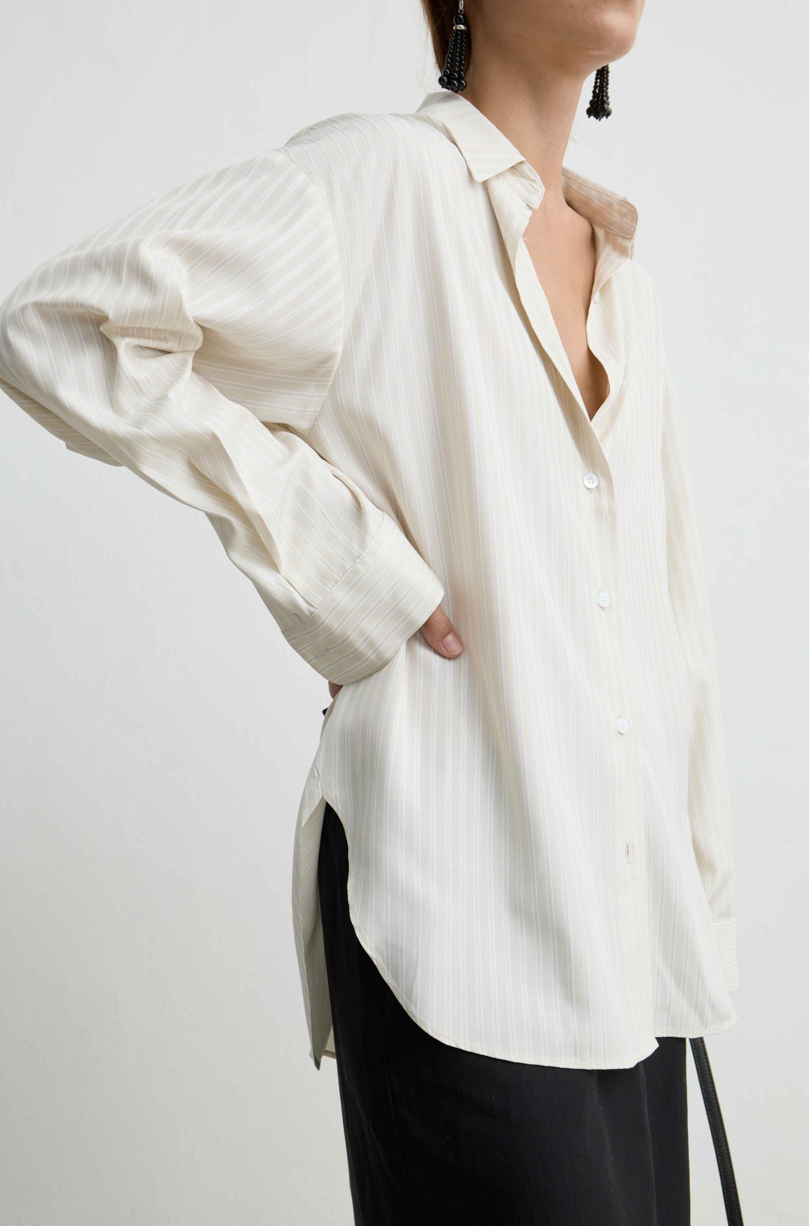 A woman wearing an oversized off-white button-up shirt with subtle vertical pinstripes and black trousers, shown from the neck down against a plain light background.
