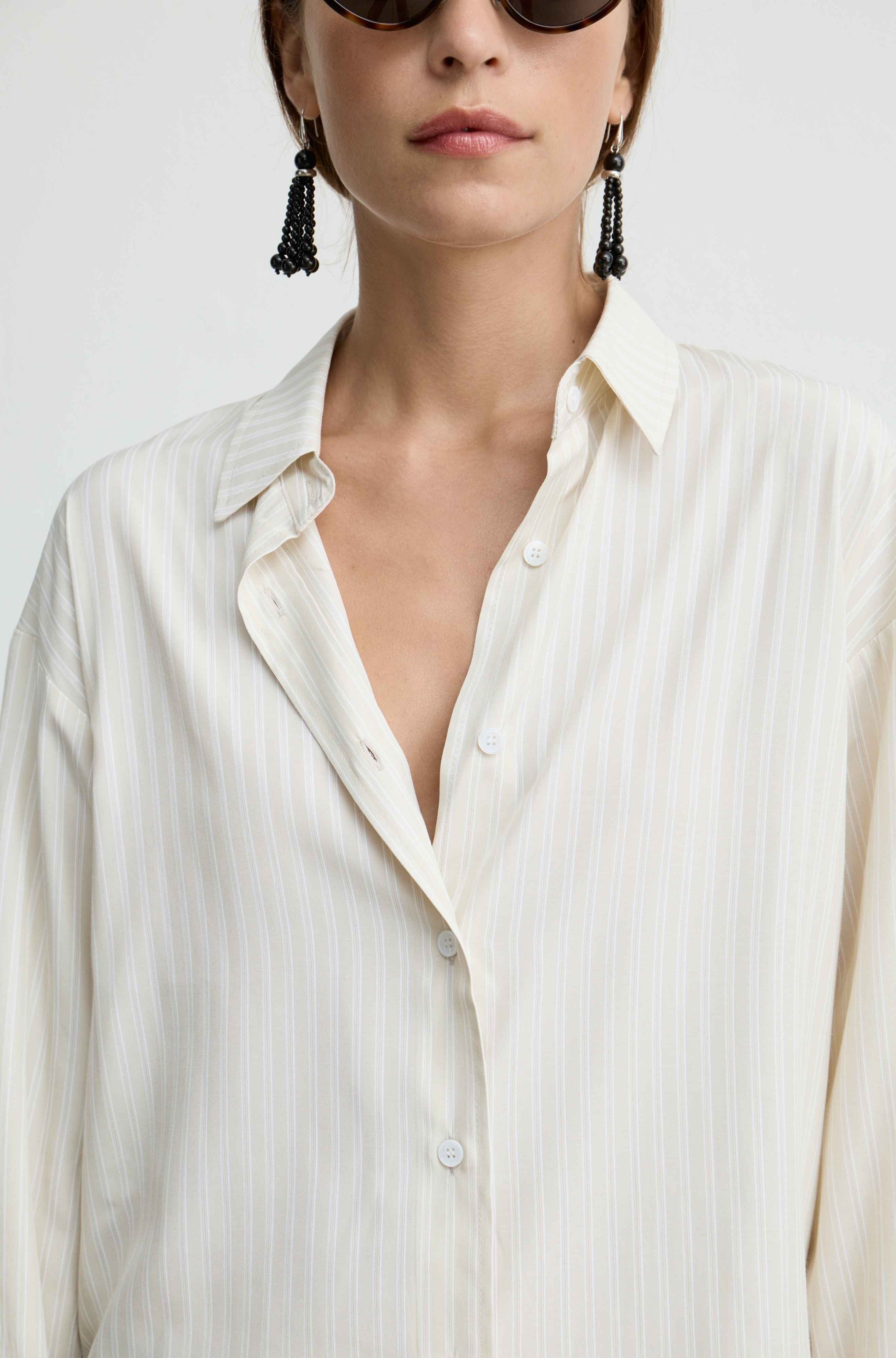 A close-up of a model wearing a cream-colored, vertically striped button-down shirt and black beaded tassel earrings against a plain light background.