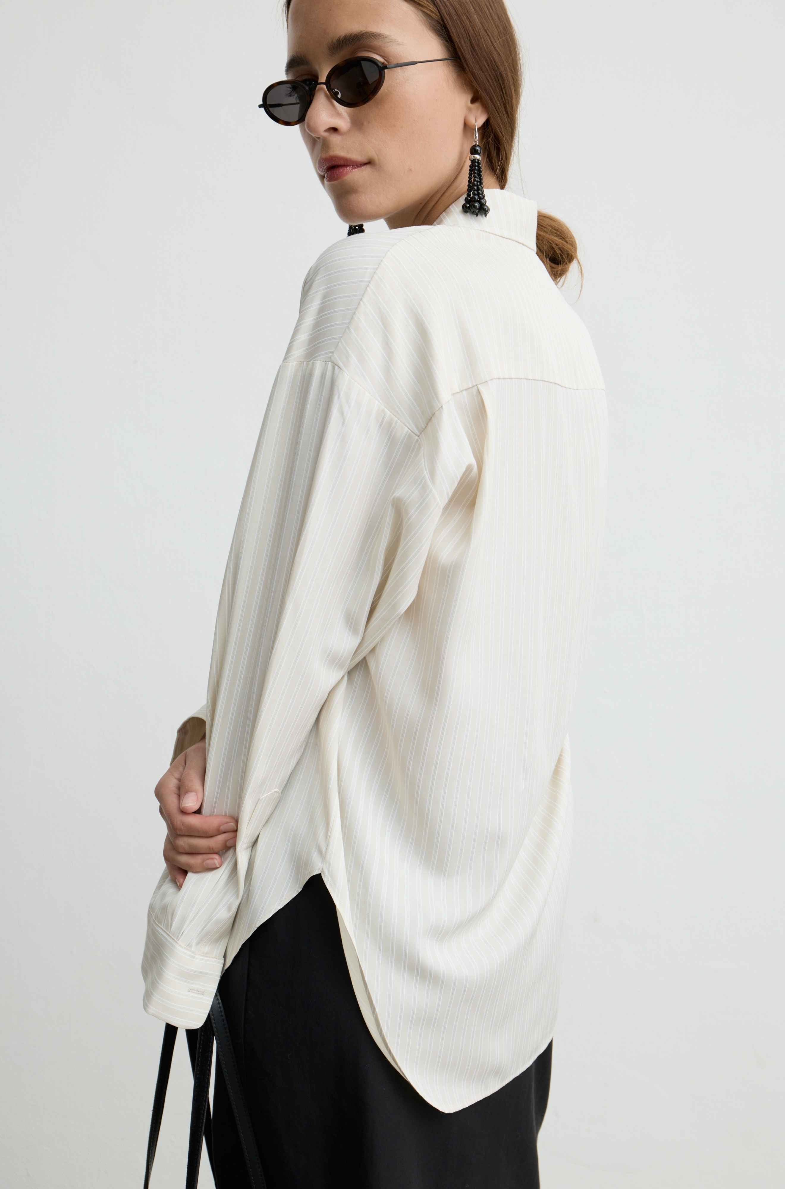A woman wearing an oversized off-white ribbed blouse with a high-low hem and black pants, photographed against a plain light gray studio background.
