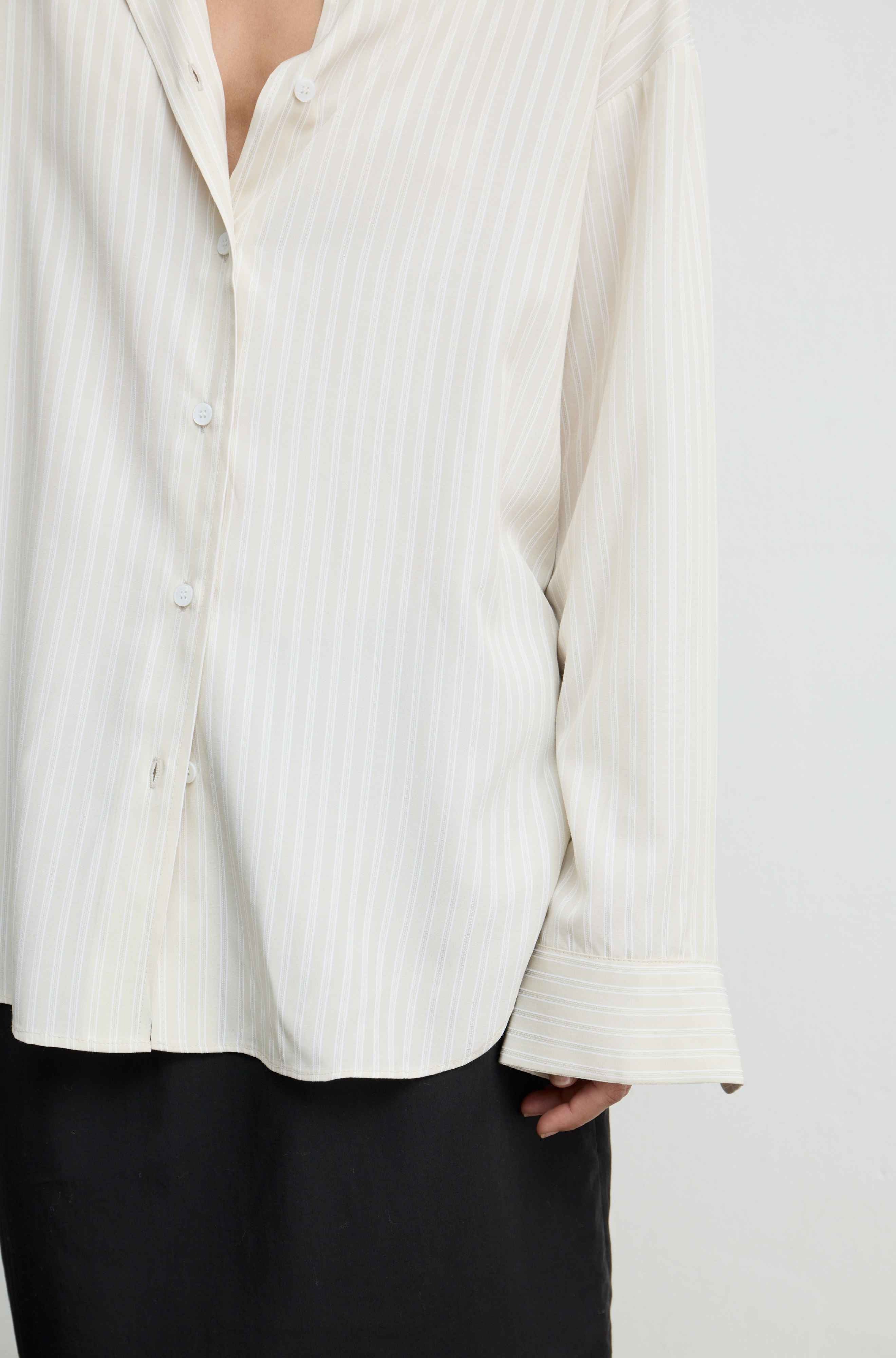 A close-up of a person wearing a cream-colored, long-sleeve button-up shirt with thin vertical stripes, paired with black trousers.