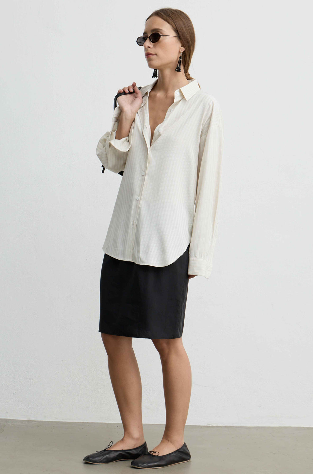 A woman modeling a cream-colored long-sleeve button-up shirt with subtle vertical pinstripes, paired with a black pencil skirt and black leather loafers, set against a plain light gray studio background.
