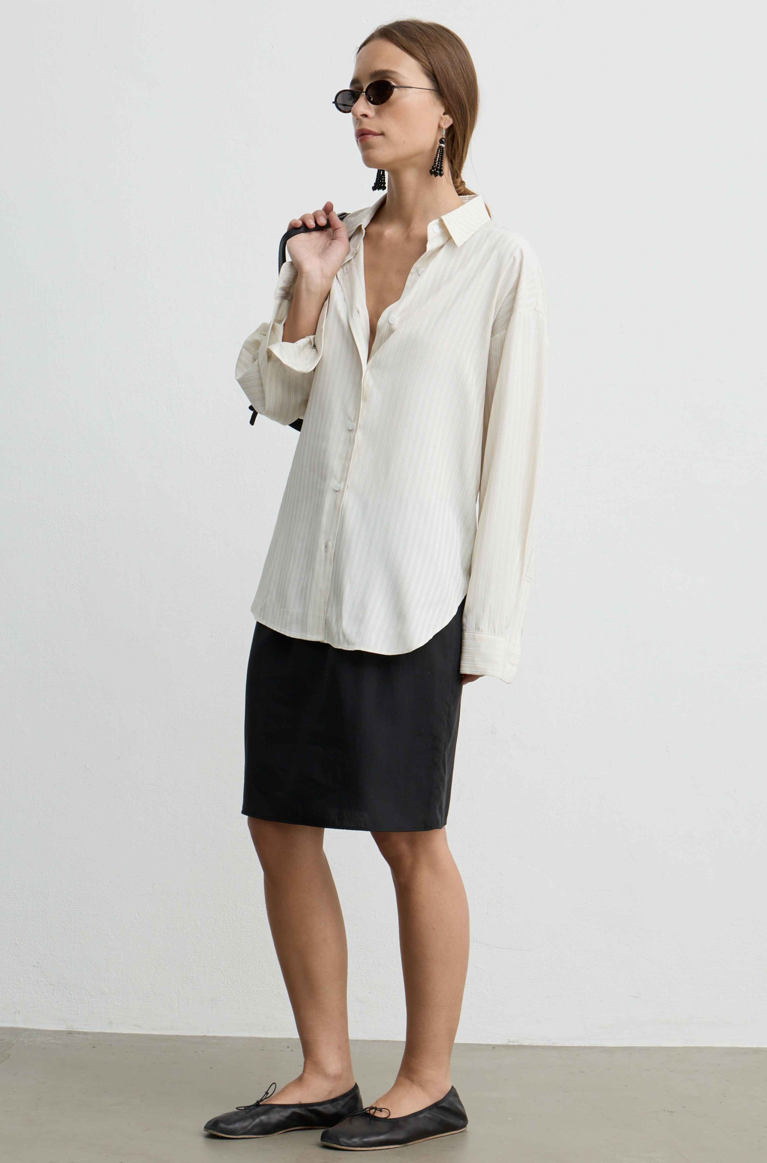 A woman modeling a cream-colored long-sleeve button-up shirt with subtle vertical pinstripes, paired with a black pencil skirt and black leather loafers, set against a plain light gray studio background.