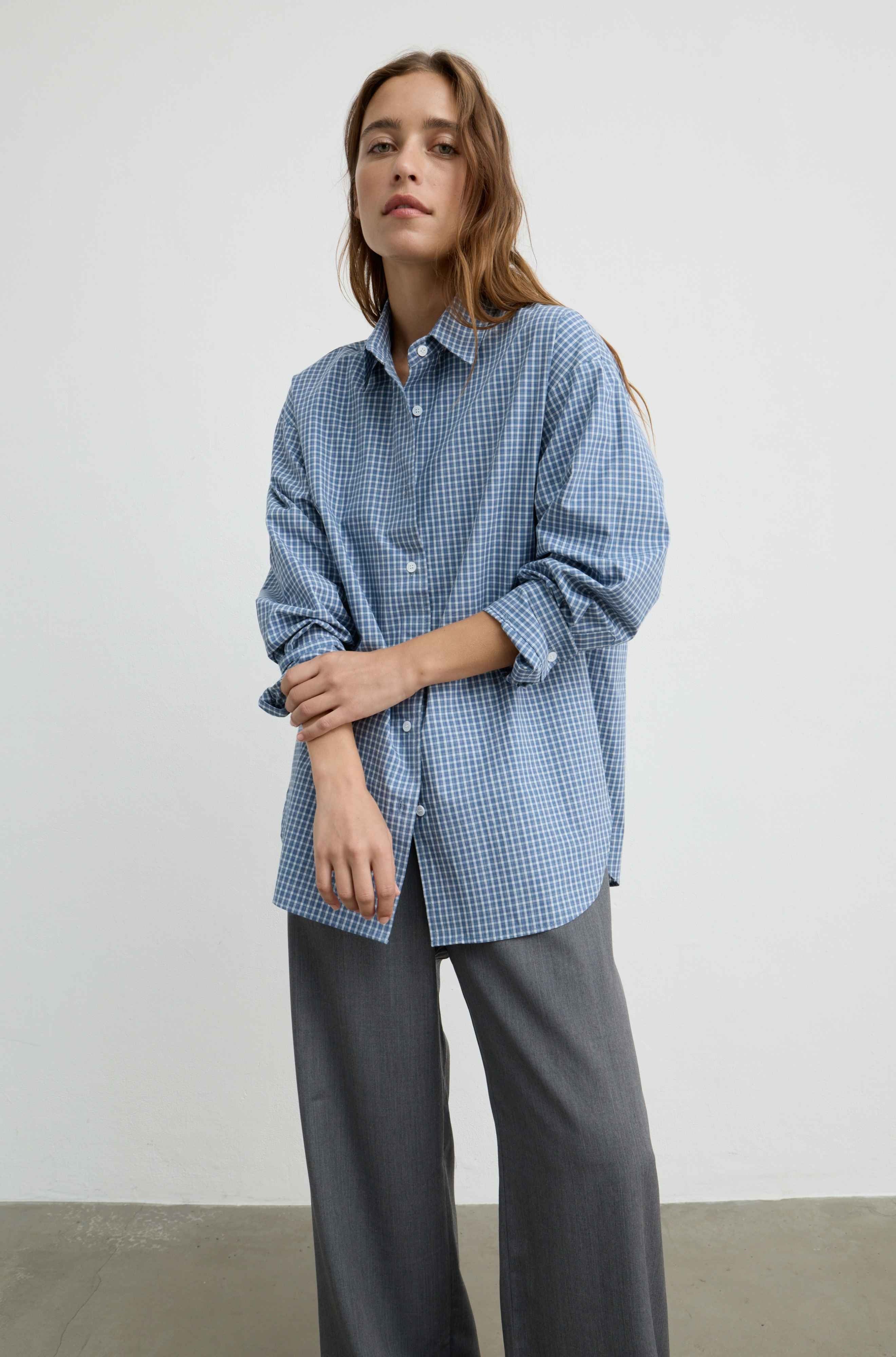 A woman modeling a light blue and white gingham button-up shirt with dark gray wide-leg trousers, standing against a plain off-white wall.