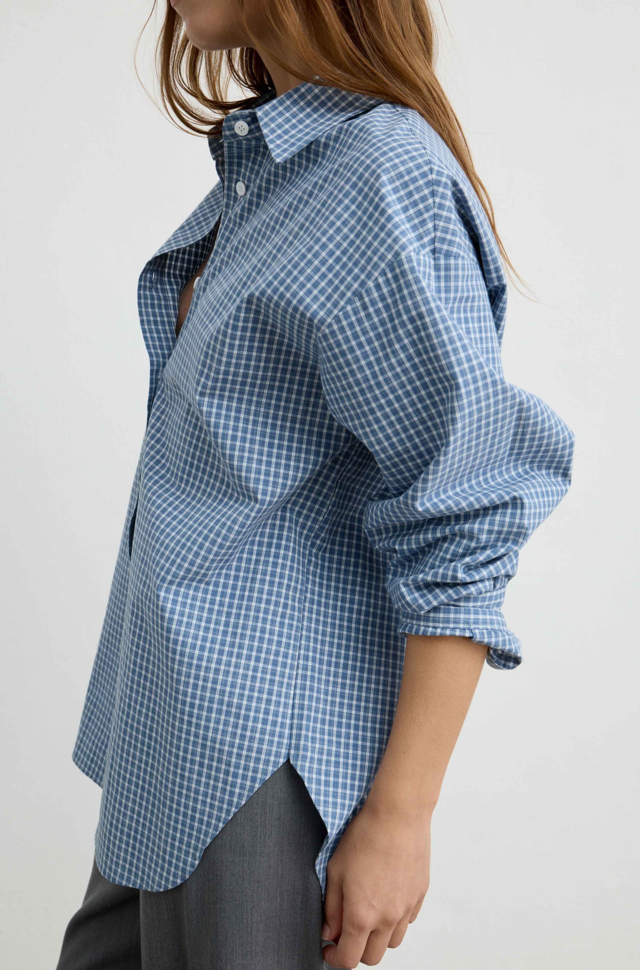 A woman wearing a blue and white gingham check long-sleeve button-up shirt, shown from the side against a plain light background.