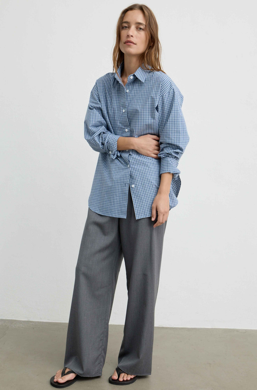A woman modeling a blue and white gingham button-up shirt tied at the waist, paired with wide-leg gray trousers and black sandals, against a plain light gray studio background.